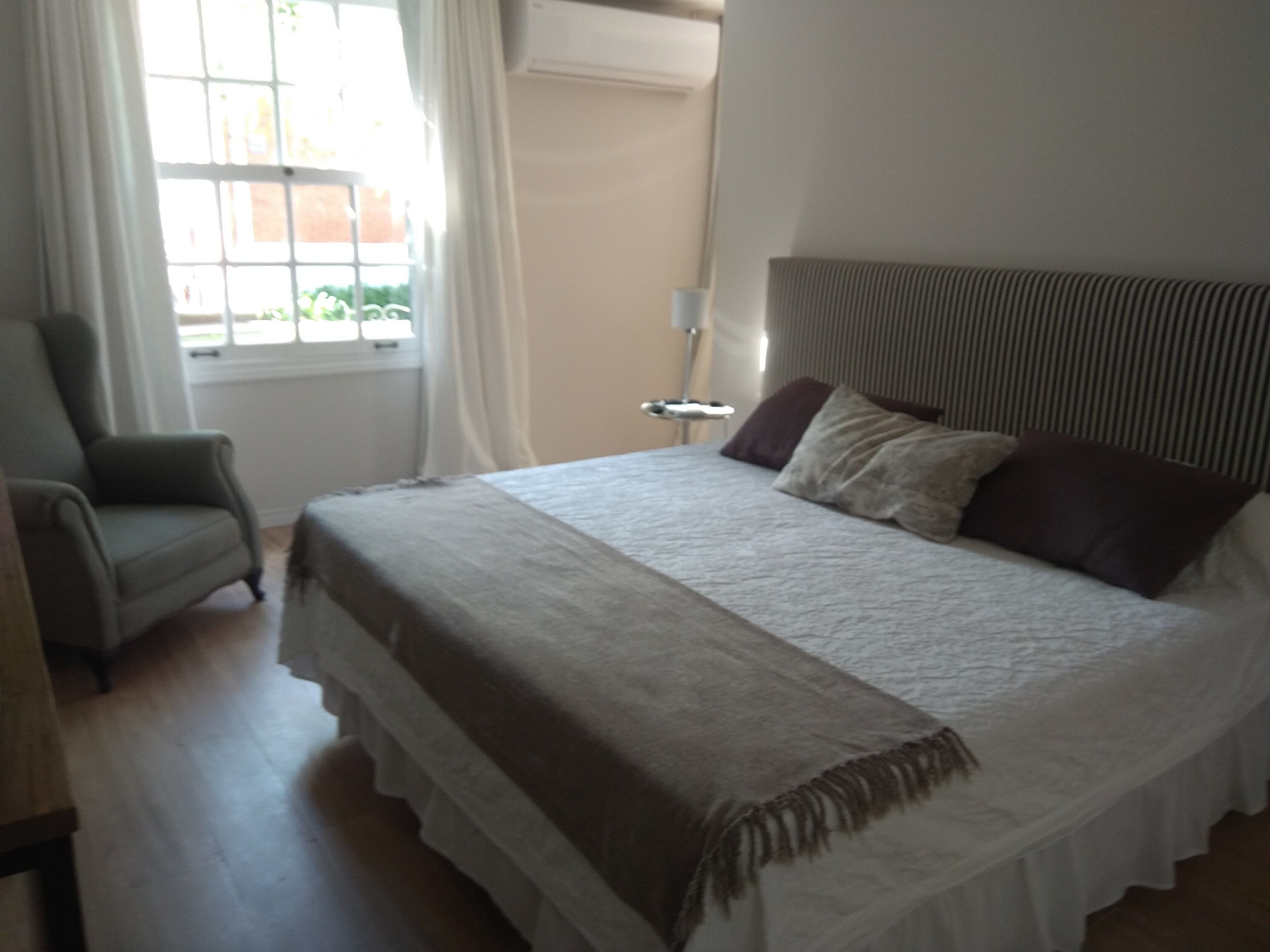 Un dormitorio con cama, una silla, una mesita de noche y una ventana con cortinas.