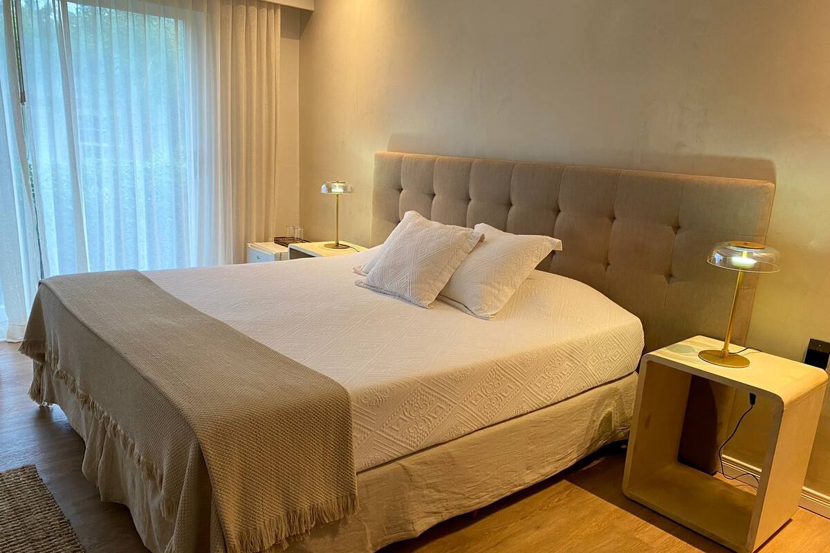 Ein ordentlich bezogenes Bett mit einer beigen Decke und Kissen, eine Nachttischlampe sowie ein kleiner Tisch mit einer Lampe.