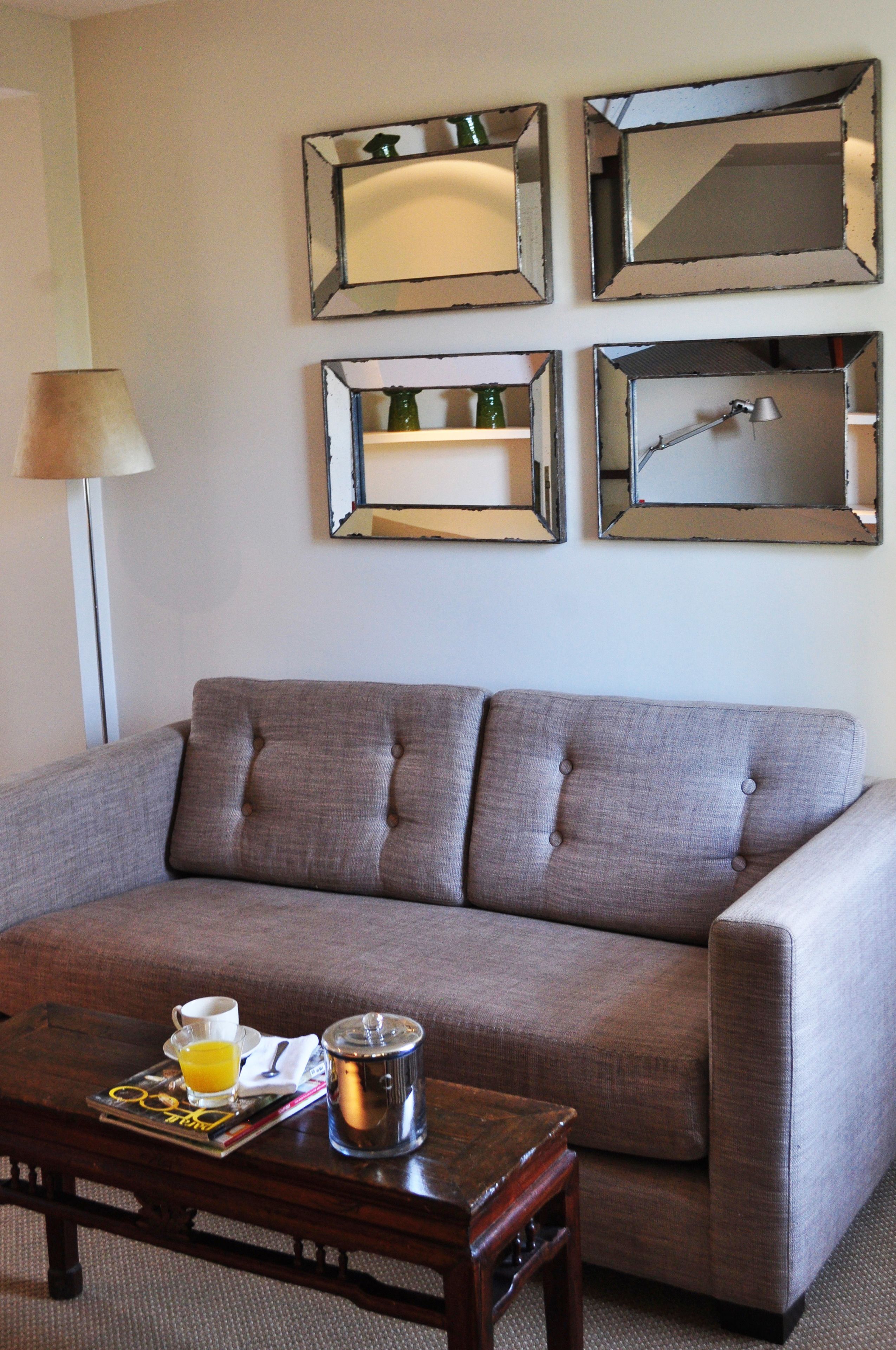 Un salón con un sofá gris, una mesa de centro de madera con una revista, una taza de café y un termo. Hay cuatro espejos en la pared.
