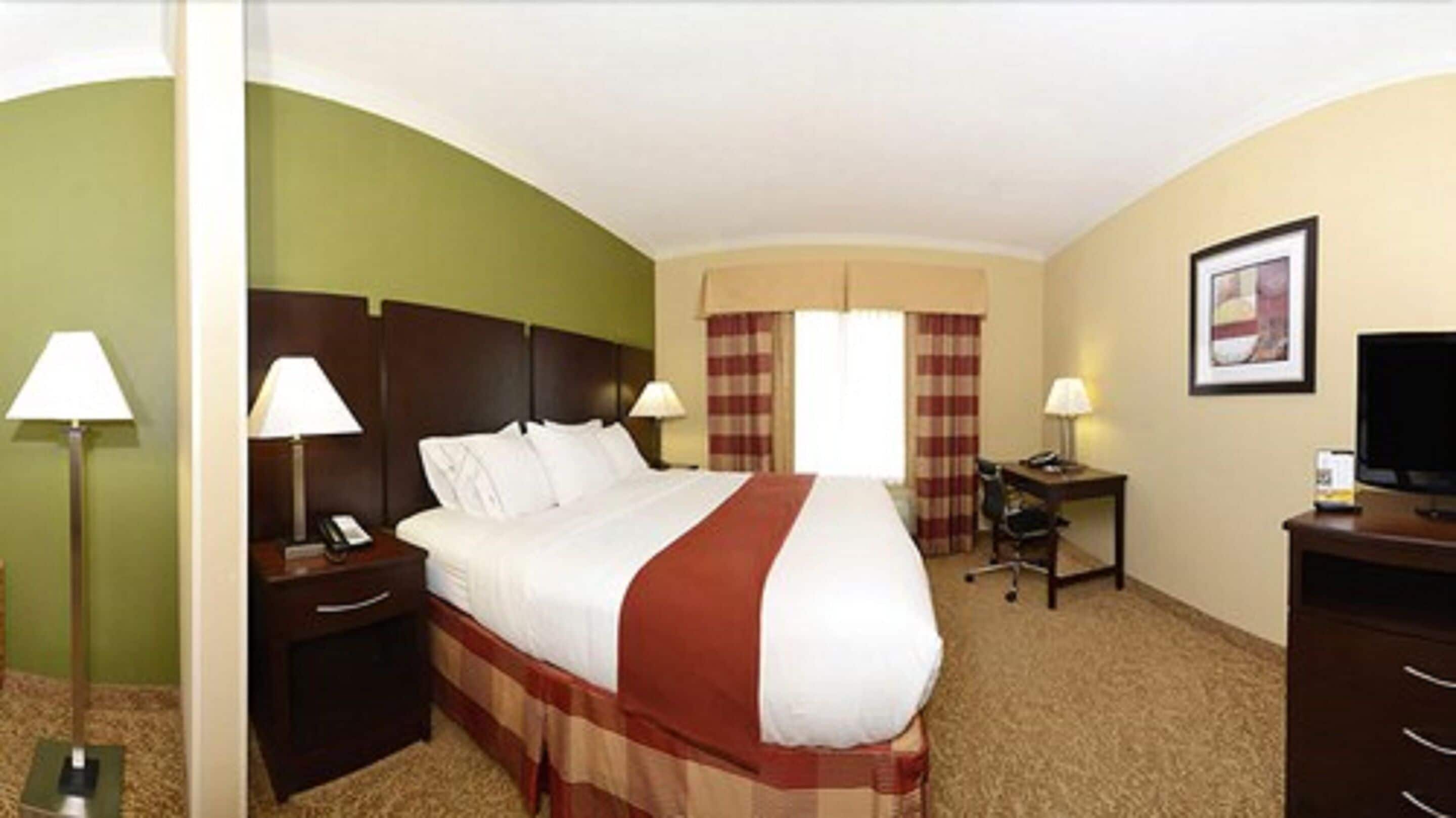 A hotel room with a large bed, two bedside tables with lamps, a desk with a chair, a television, and a framed picture on the wall.