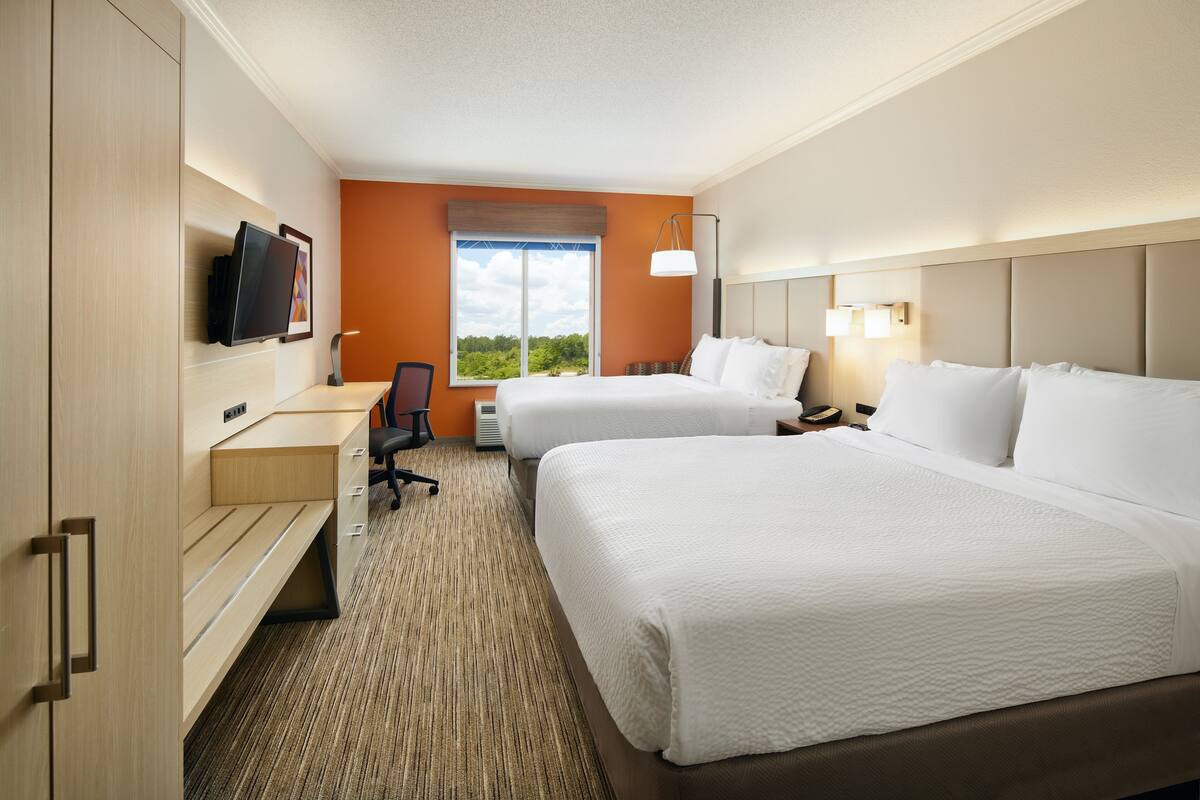 A hotel room with two beds, a desk, a chair, a TV, and a window with a view of greenery.
