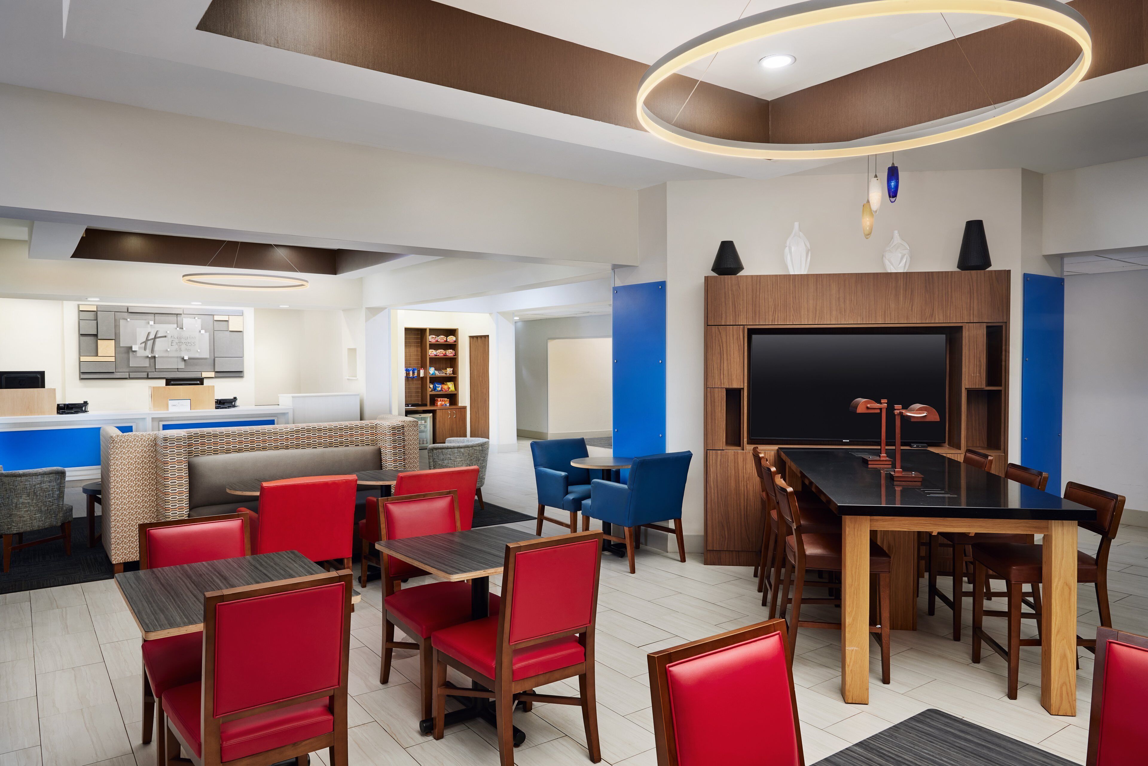 A modern hotel lobby with a dining area, a reception desk, and a large TV screen.