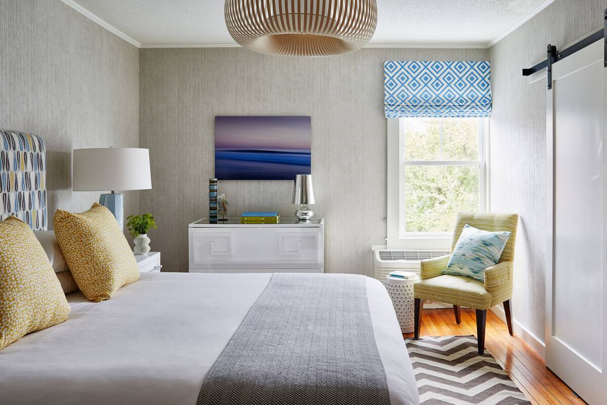 Un dormitorio ordenado con cama, mesita de noche, lámpara y una silla junto a la ventana.