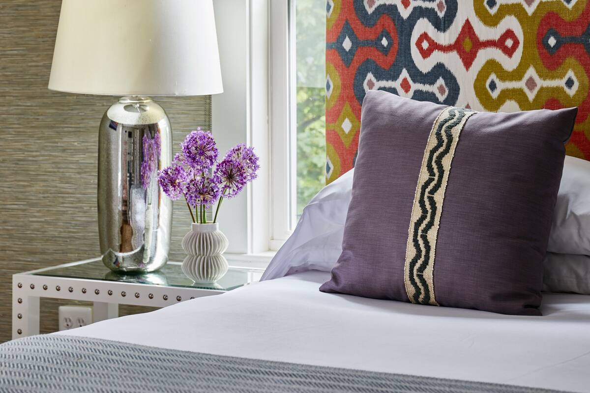 Un dormitorio ordenado con una cama, una mesita de noche con lámpara, un jarrón con flores y una ventana con cortina estampada.