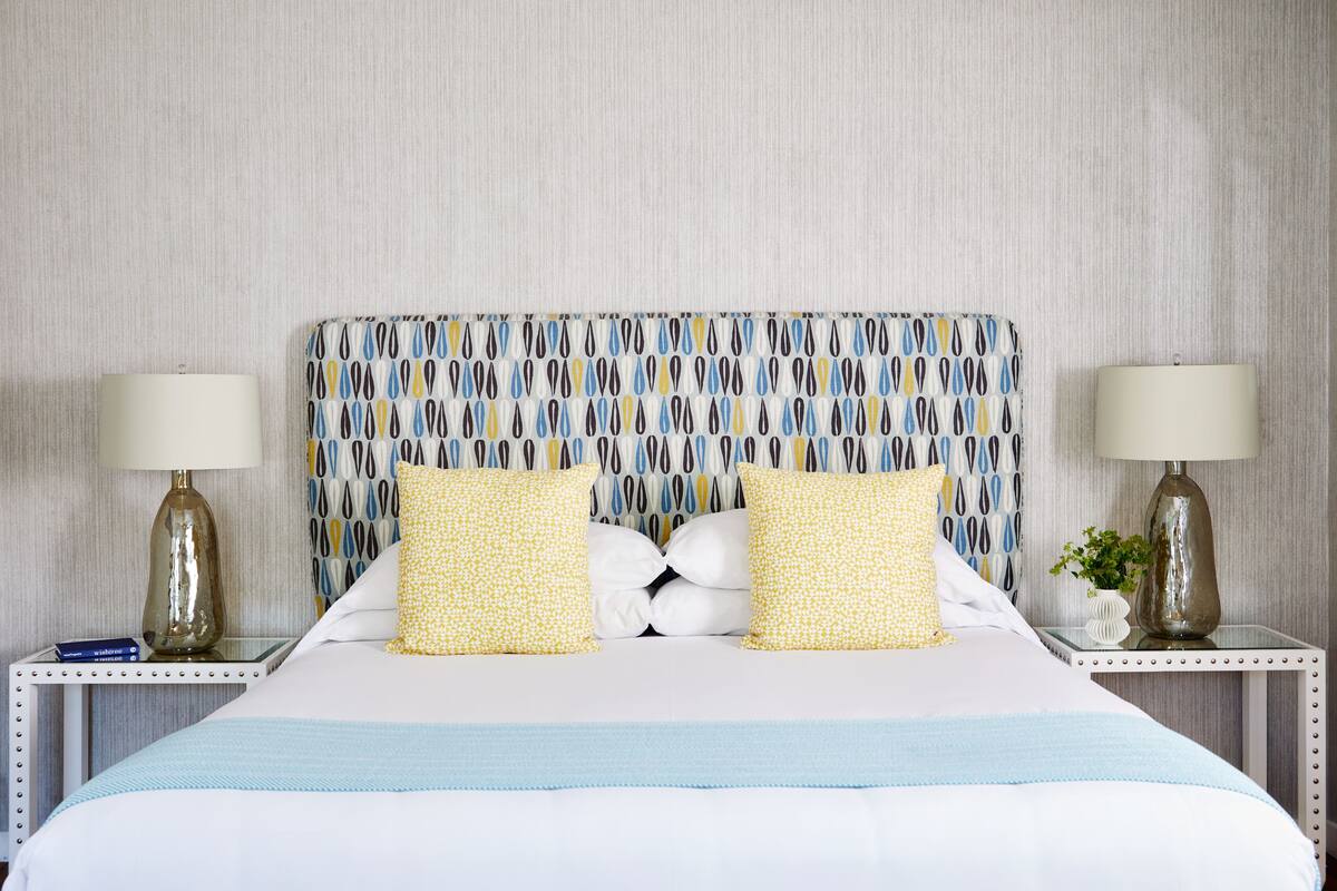 Una cama bien hecha con un cabecero estampado, dos mesitas de noche con lámparas y una colcha azul a rayas.