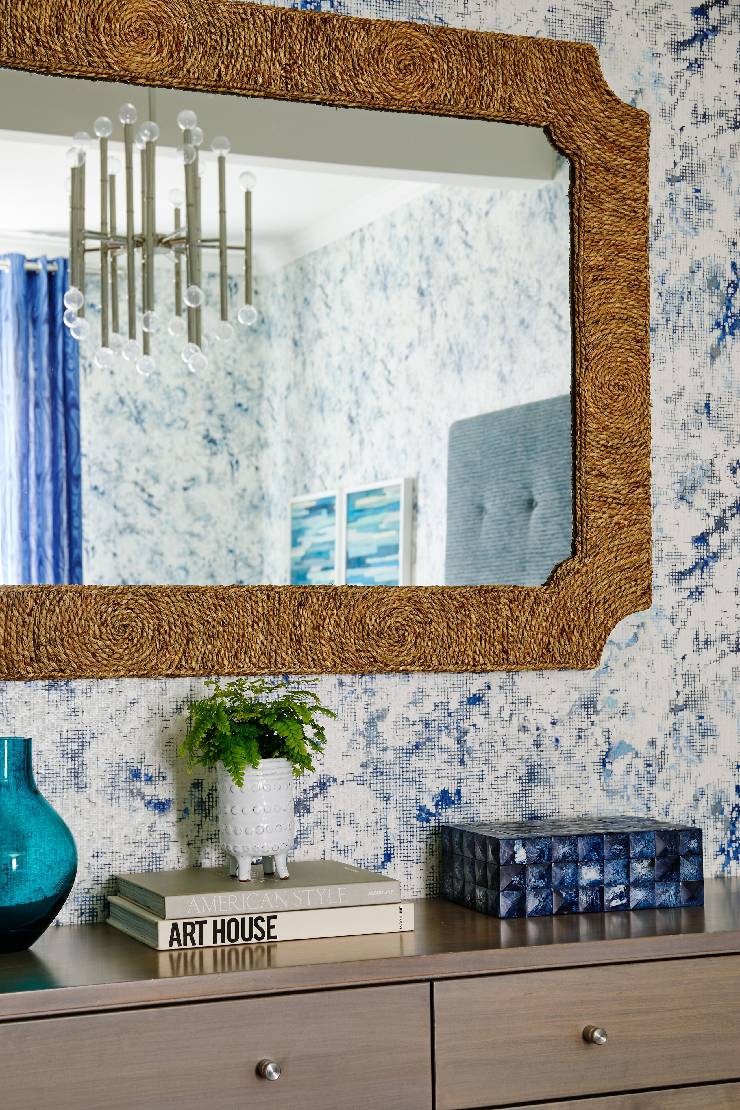 A mirror with a woven frame, a blue vase, a potted plant, and a book titled "Art House" on a wooden dresser against a blue and white patterned wall.