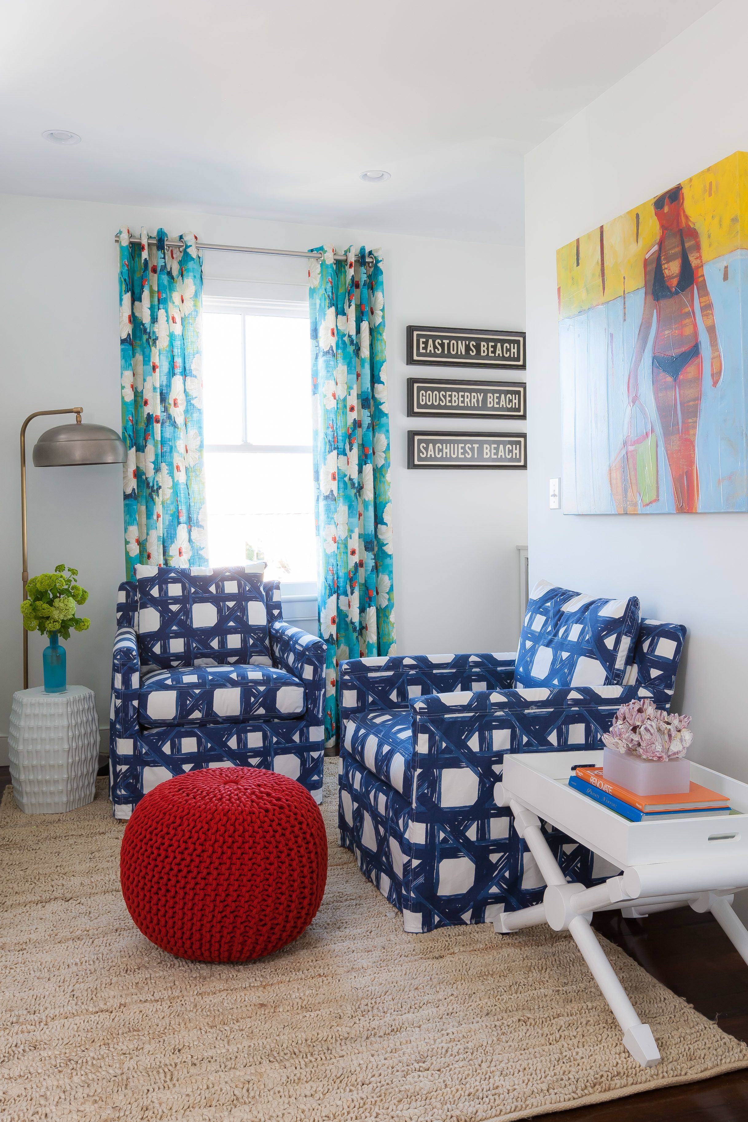 A living room with patterned armchairs, a red ottoman, and a white side table with books.