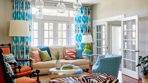 A living room with a sofa, armchairs, a coffee table, and a lamp. There are curtains, a door, and a window.