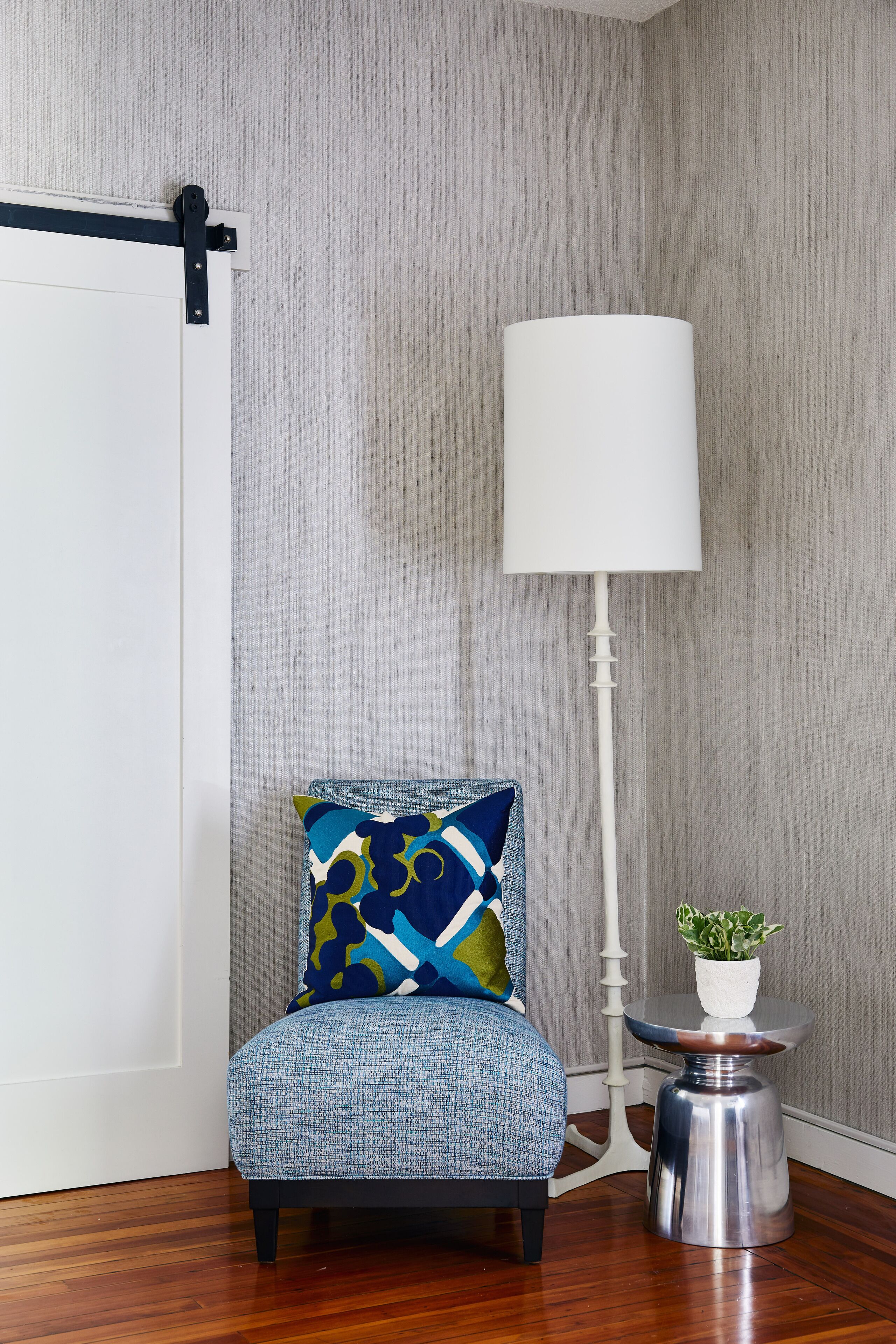 Une chaise avec un coussin à motifs, un lampadaire blanc et une petite table sur laquelle est posée une plante en pot.