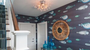 A hallway with a dark wall featuring fish patterns, a wicker fan, and blue glass bottles on a metal table.