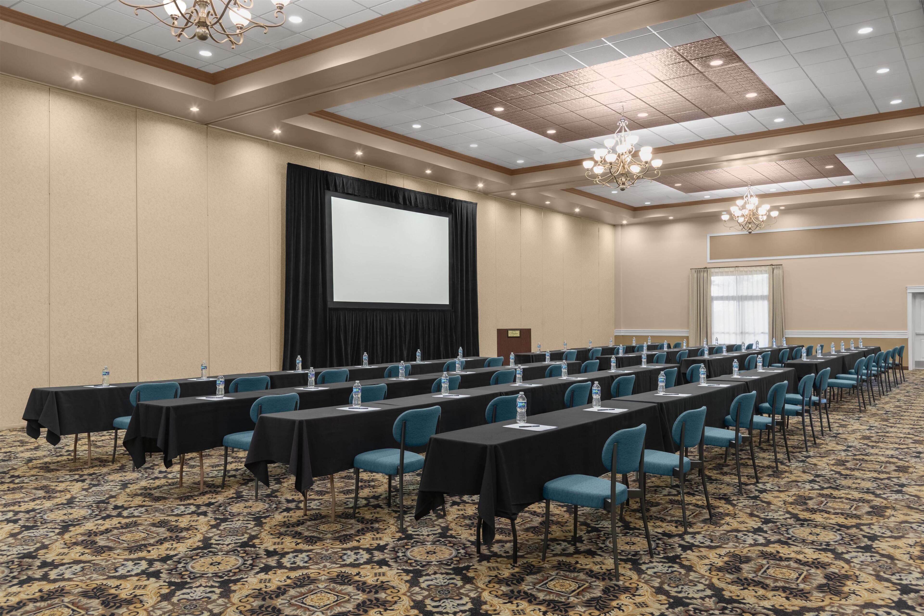 A conference room with long tables, chairs, a projector screen, and a carpeted floor.