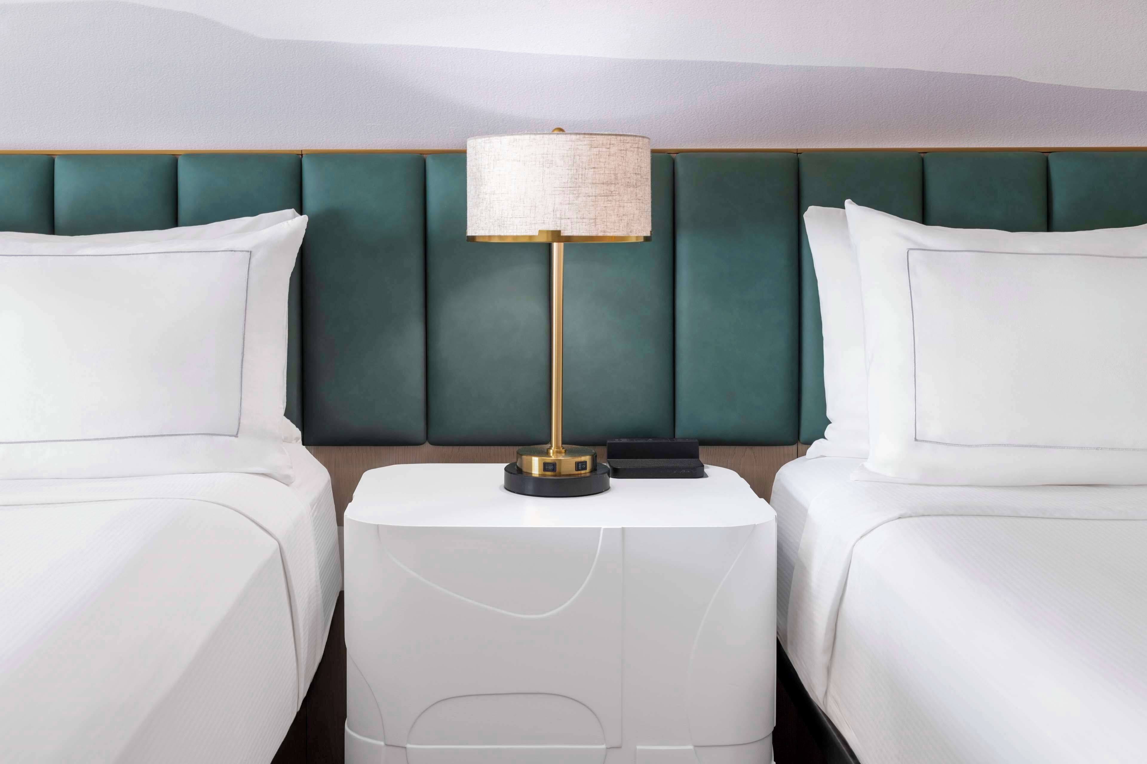 A hotel room with two beds, a green upholstered headboard, a white bedside table with a lamp, and a white pillow.