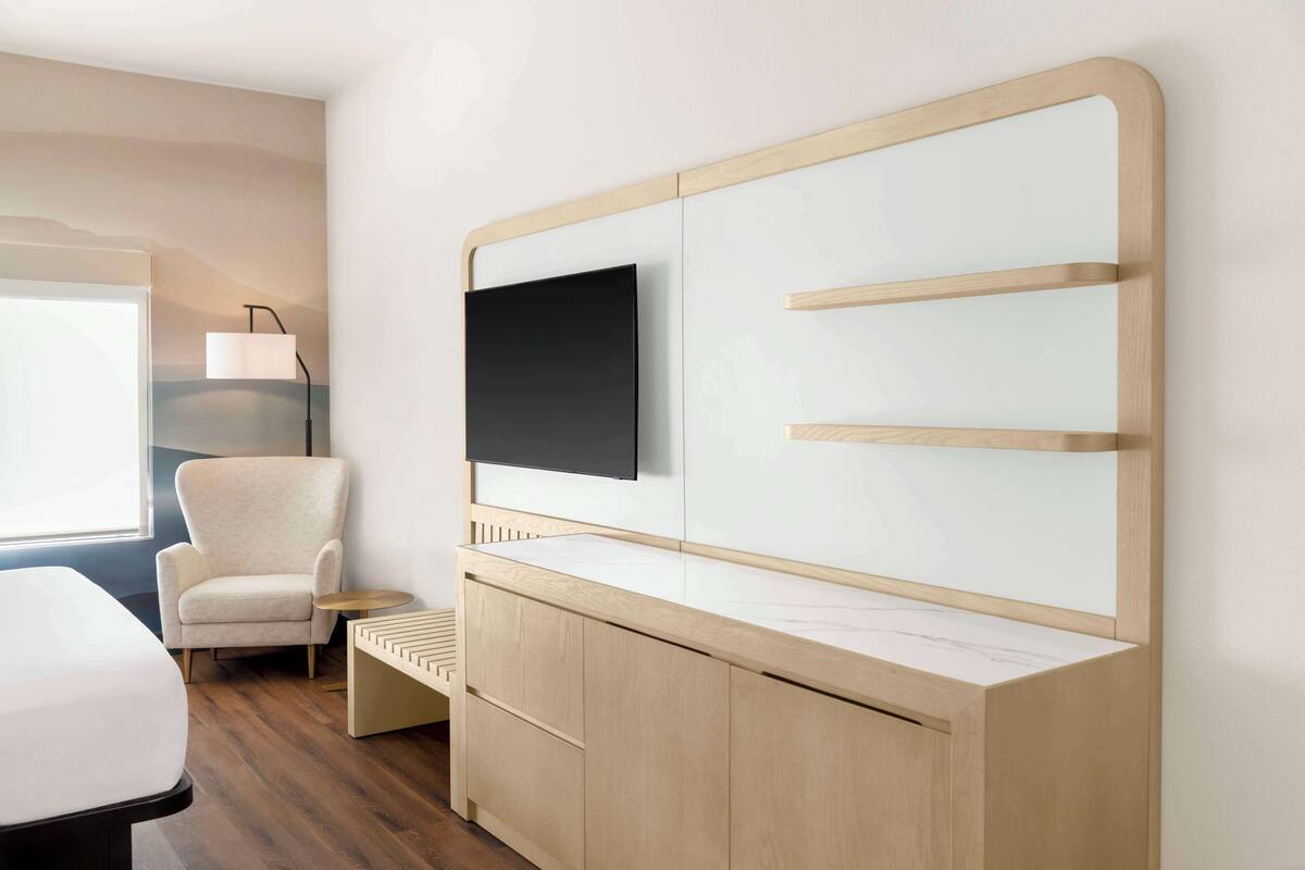 A modern hotel room with a large flat-screen TV mounted on a wooden cabinet, a comfortable armchair, and a small side table.