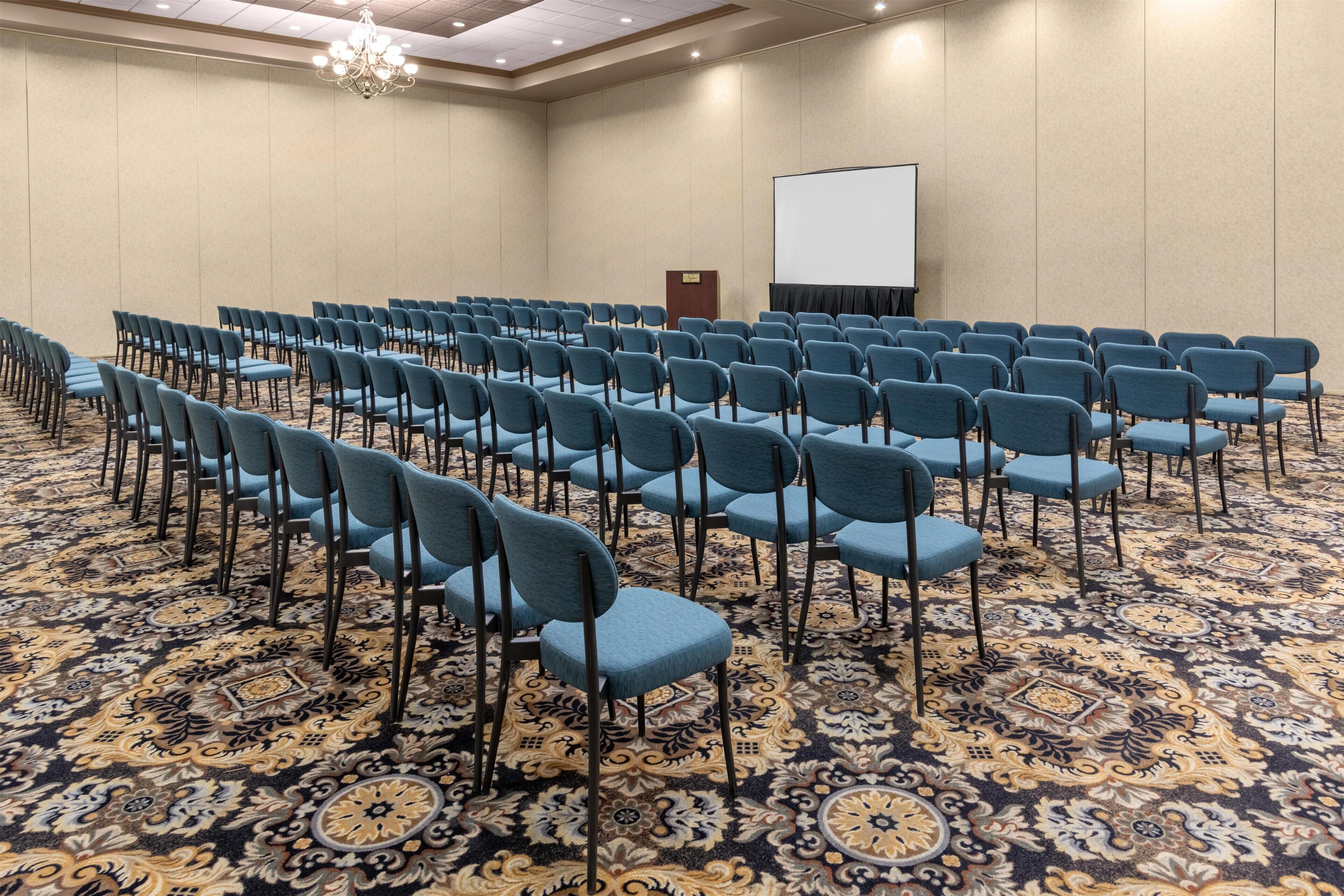 A conference room with rows of chairs, a projector screen, and a carpeted floor.