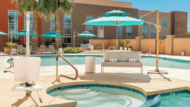 Una piscina con sillas de descanso y sombrillas, un jacuzzi y un edificio al fondo.