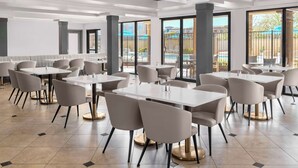 Un comedor moderno con mesas y sillas blancas, ventanas amplias y vistas a la piscina.