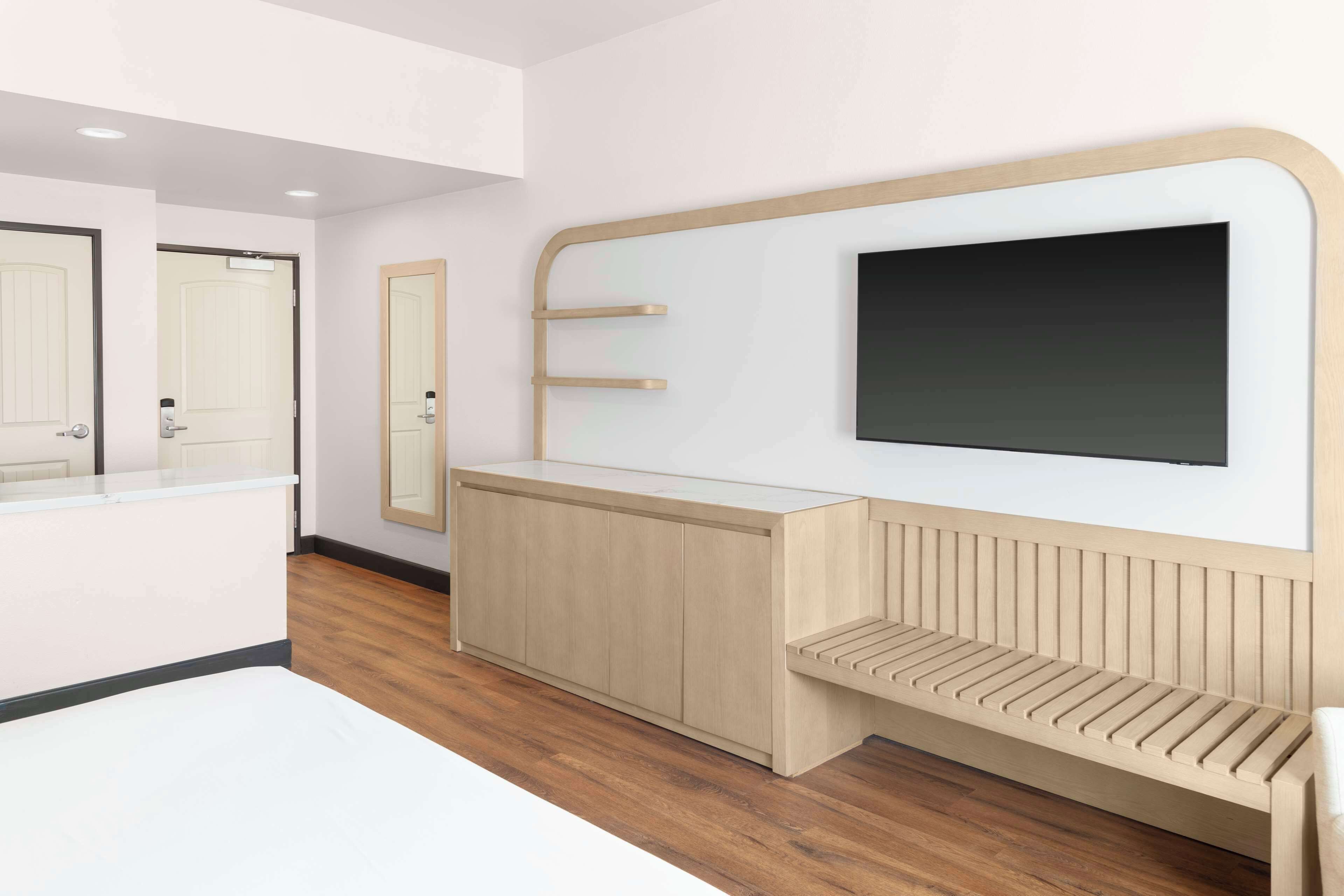 Ein modernes Hotelzimmer mit Holzboden, einem weißen Bett, einer Holzbank, einem großen Spiegel und einem schwarzen Fernseher an der Wand.