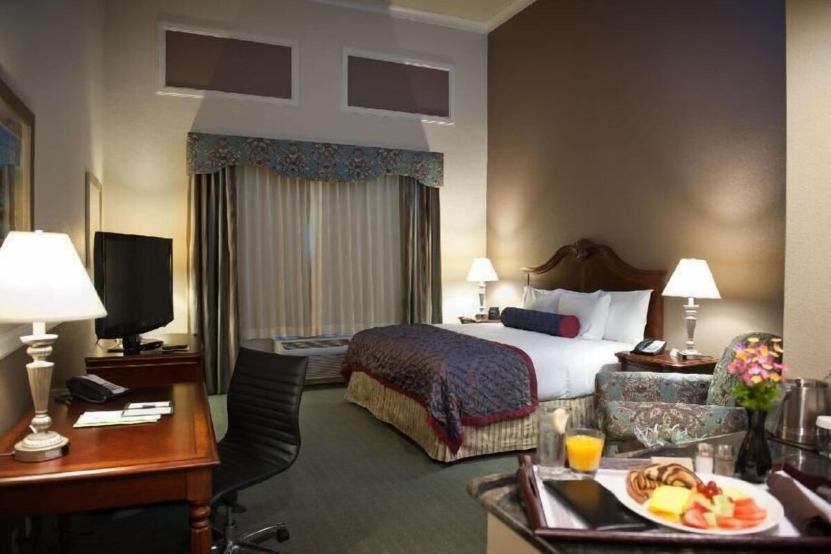 Habitación de hotel con cama, dos lámparas de noche, escritorio con lámpara, televisor, silla, mesa de comedor con desayuno y ventana con cortinas.