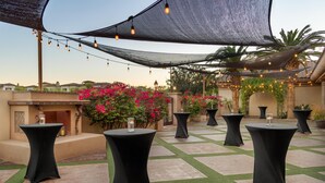 Un patio al aire libre con mesas redondas, luces colgantes y un fondo de palmeras y flores.