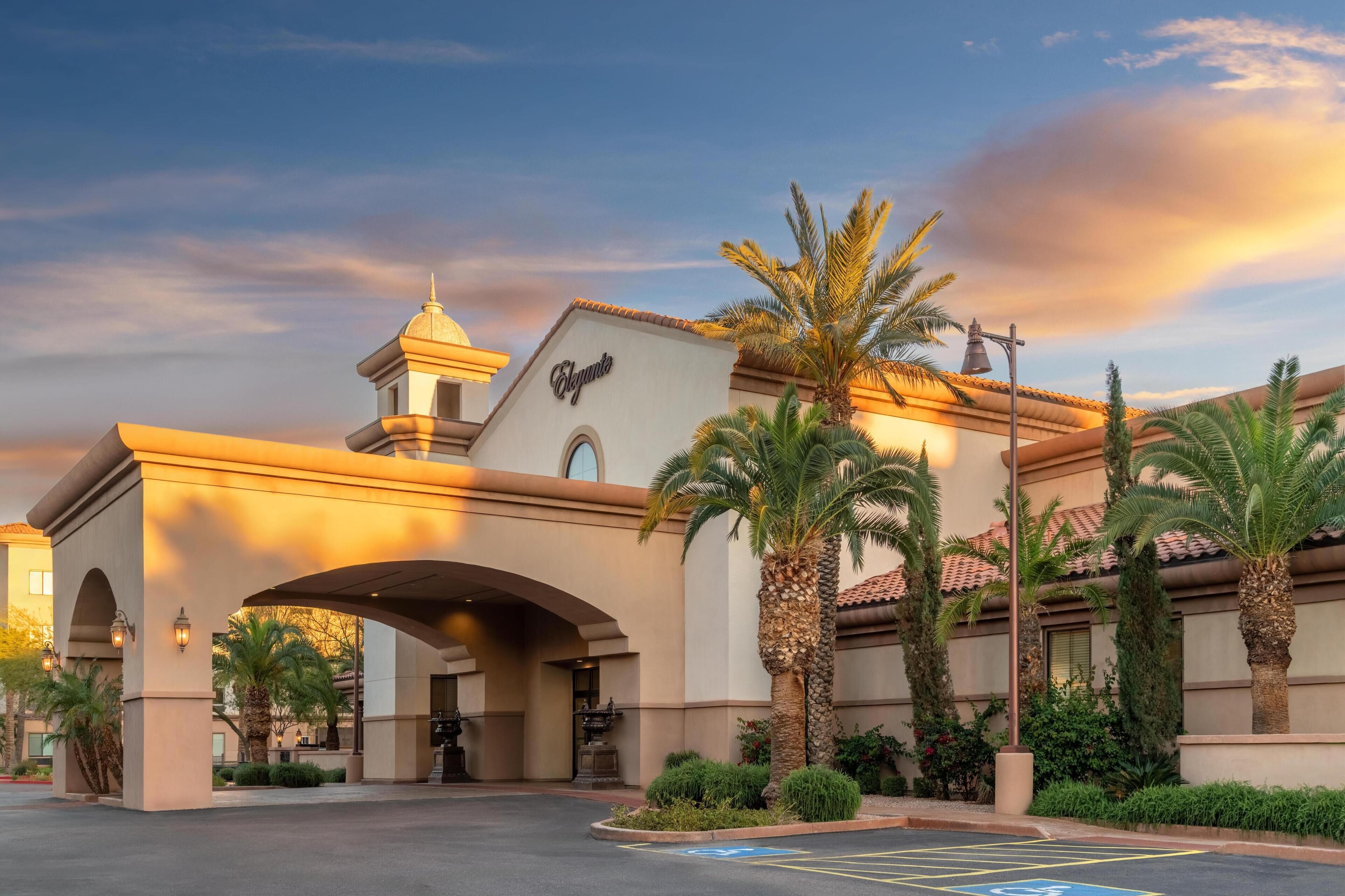 Entrada de hotel con un camino cubierto, palmeras y una torre de campanas.