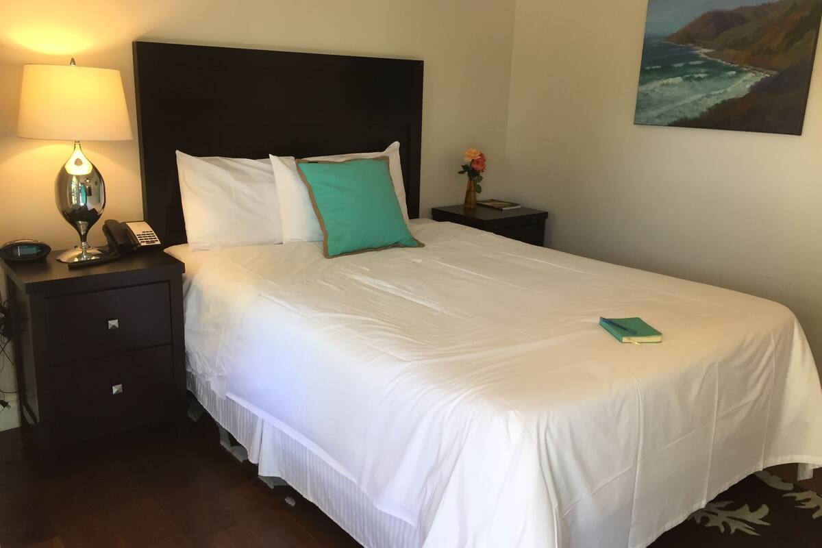 A neatly made bed with white bedding and a teal pillow, a bedside lamp, a phone, and a small table with a vase of flowers.
