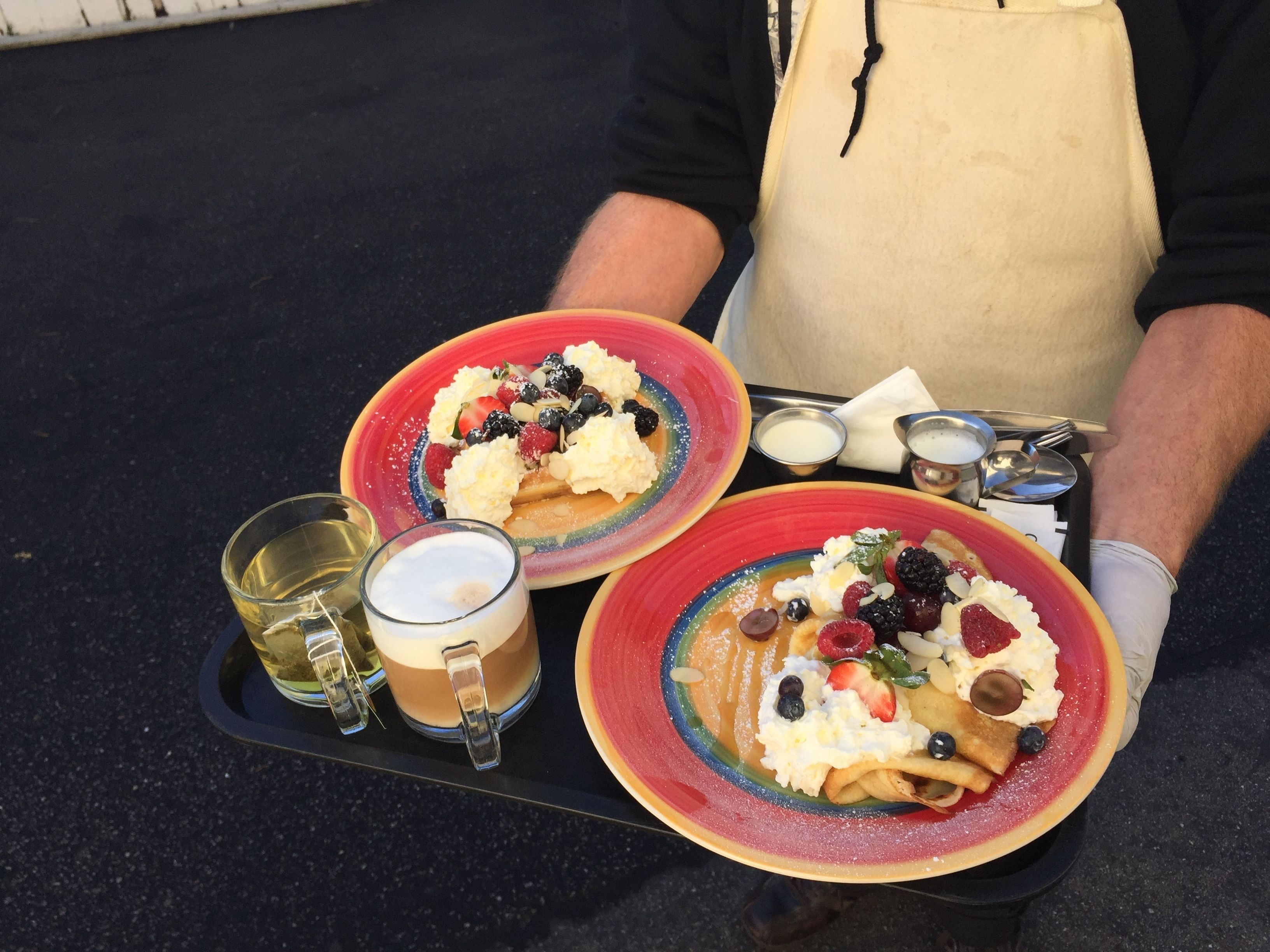 Una persona serve due piatti di pancake con frutta e panna montata, un bicchiere di caffè e un piccolo contenitore di sciroppo.