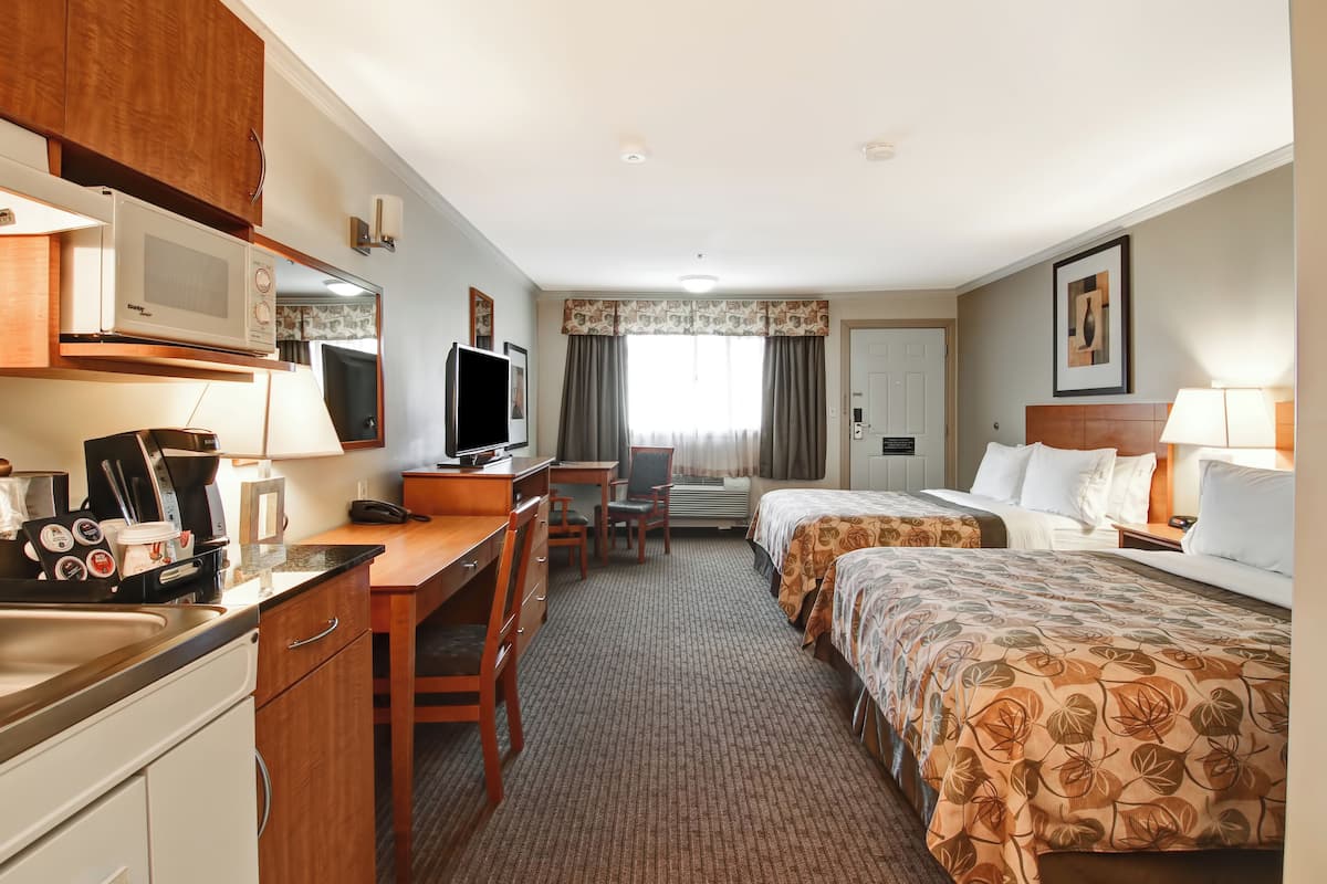 A hotel room with a kitchenette, a desk with a chair, two beds with floral bedspreads, a television, and a window with curtains.
