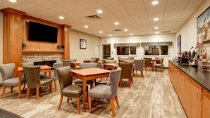 A spacious dining area with multiple tables and chairs, a stone fireplace, and a television mounted on the wall. - Canalta Hotel Jurassic – Drumheller (Drumheller)