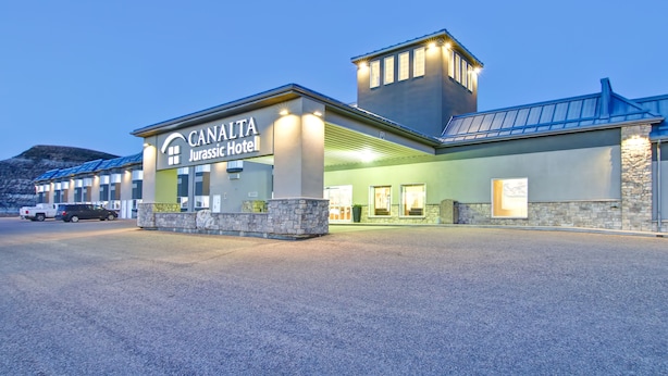 The exterior of the Canalta Jurassic Hotel with a prominent sign, stone facade, and a parking lot with vehicles. - Canalta Hotel Jurassic – Drumheller (Drumheller)