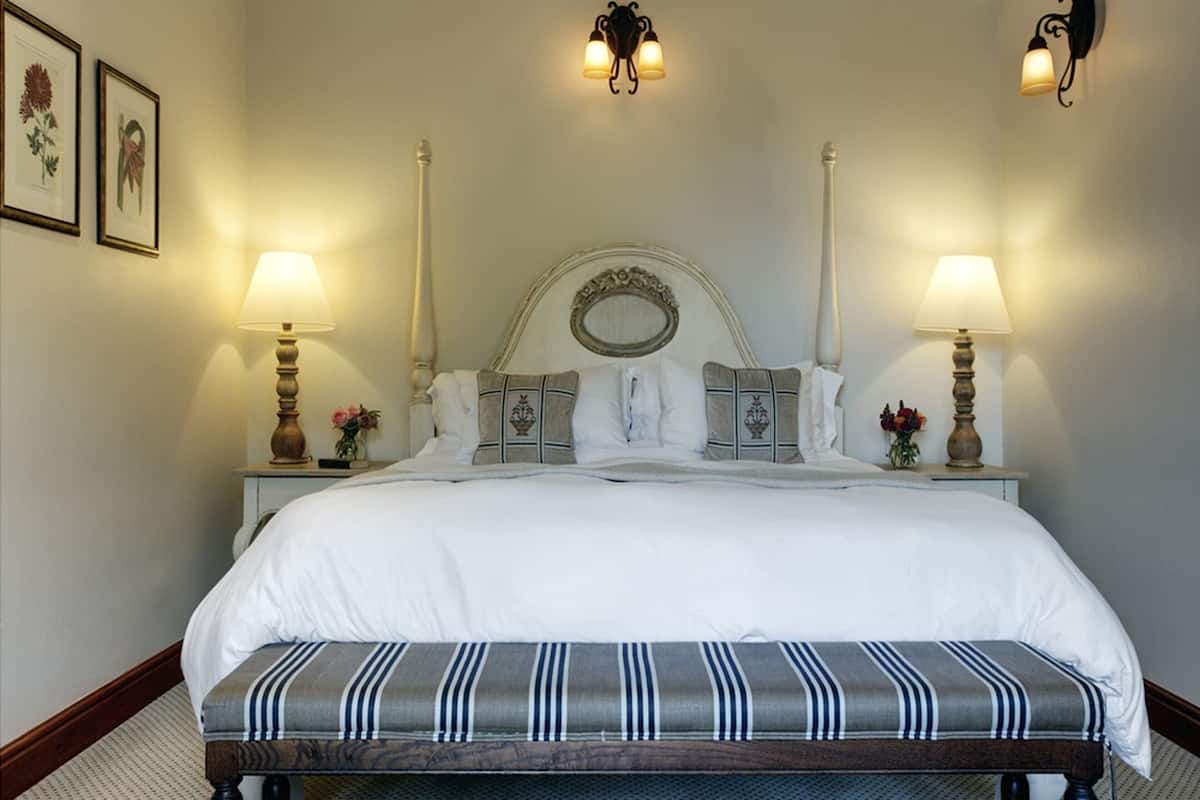 A neatly made bed with a white comforter and striped bench, flanked by two bedside tables with lamps and framed pictures on the wall.