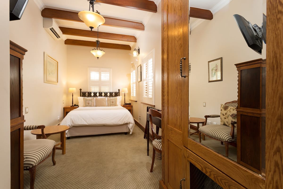 A hotel room with a bed, a wooden wardrobe, a chair, a small table, and a mirror.