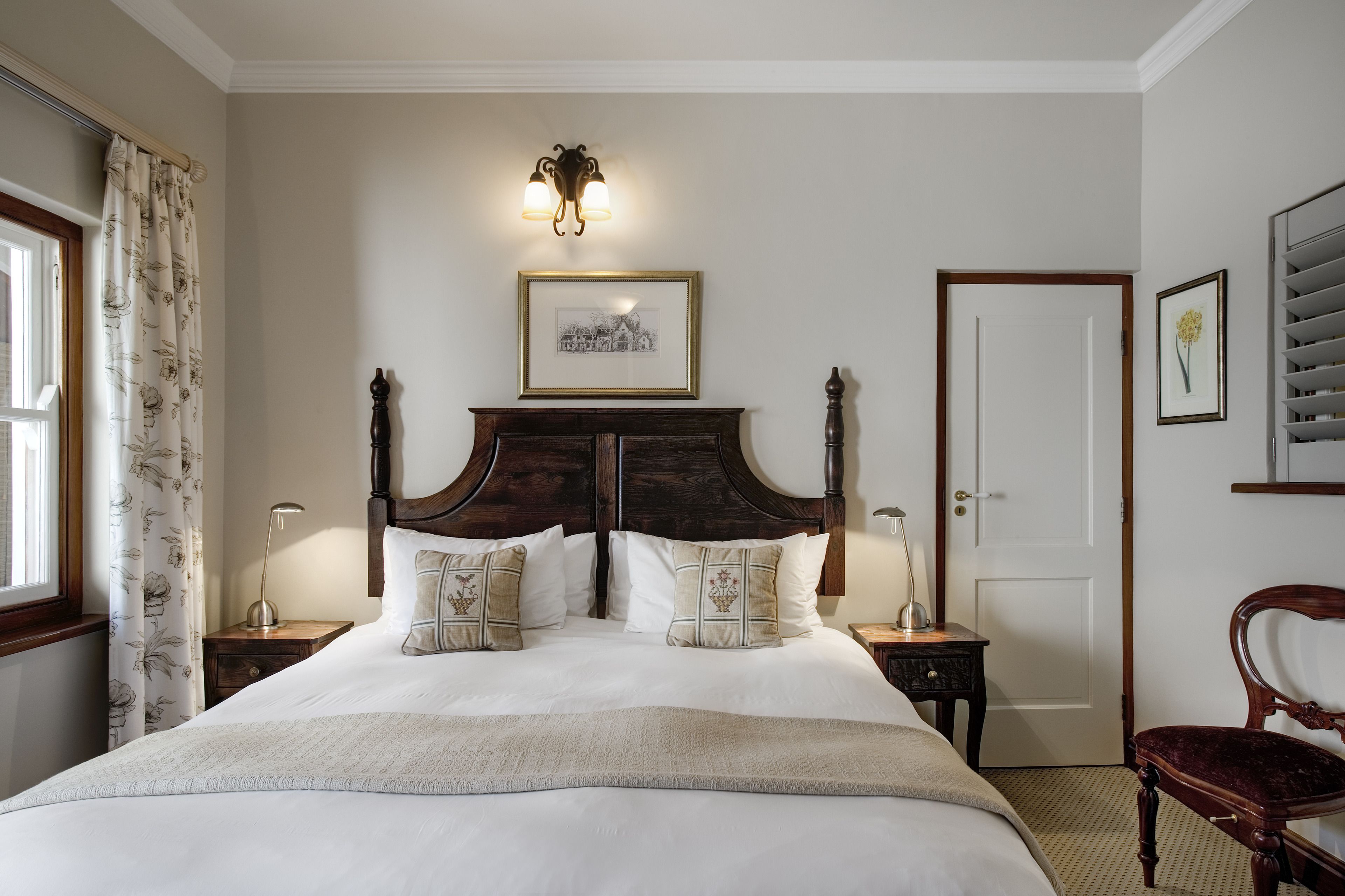 A neatly made bed with a headboard, two bedside tables with lamps, a framed picture above the headboard, a door, and a chair.