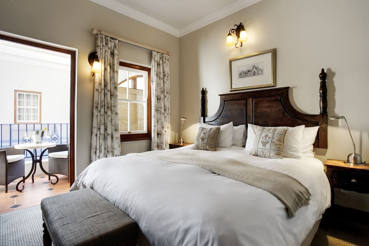 A bedroom with a large bed, a wooden headboard, a bedside table with a lamp, and a balcony with a table and chairs.