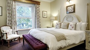 A bedroom with a large bed, a chair, a bench, a window with curtains, a nightstand, and a wall-mounted lamp. - Coopmanhuijs Boutique Hotel (Stellenbosch)