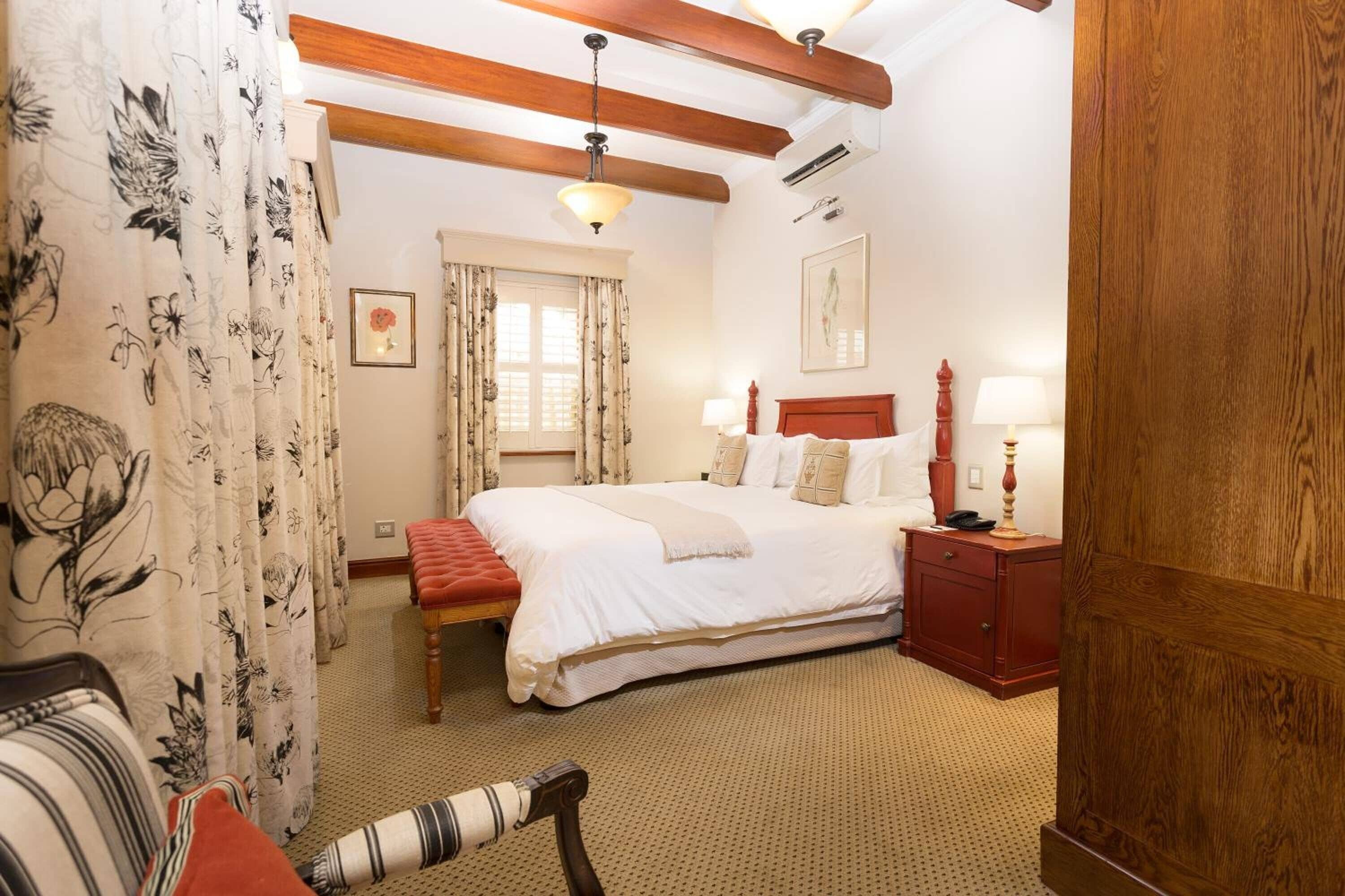 Un dormitorio con una cama grande, cabecera de madera, una mesita de noche con lámpara, una ventana con cortinas y un banco de madera.