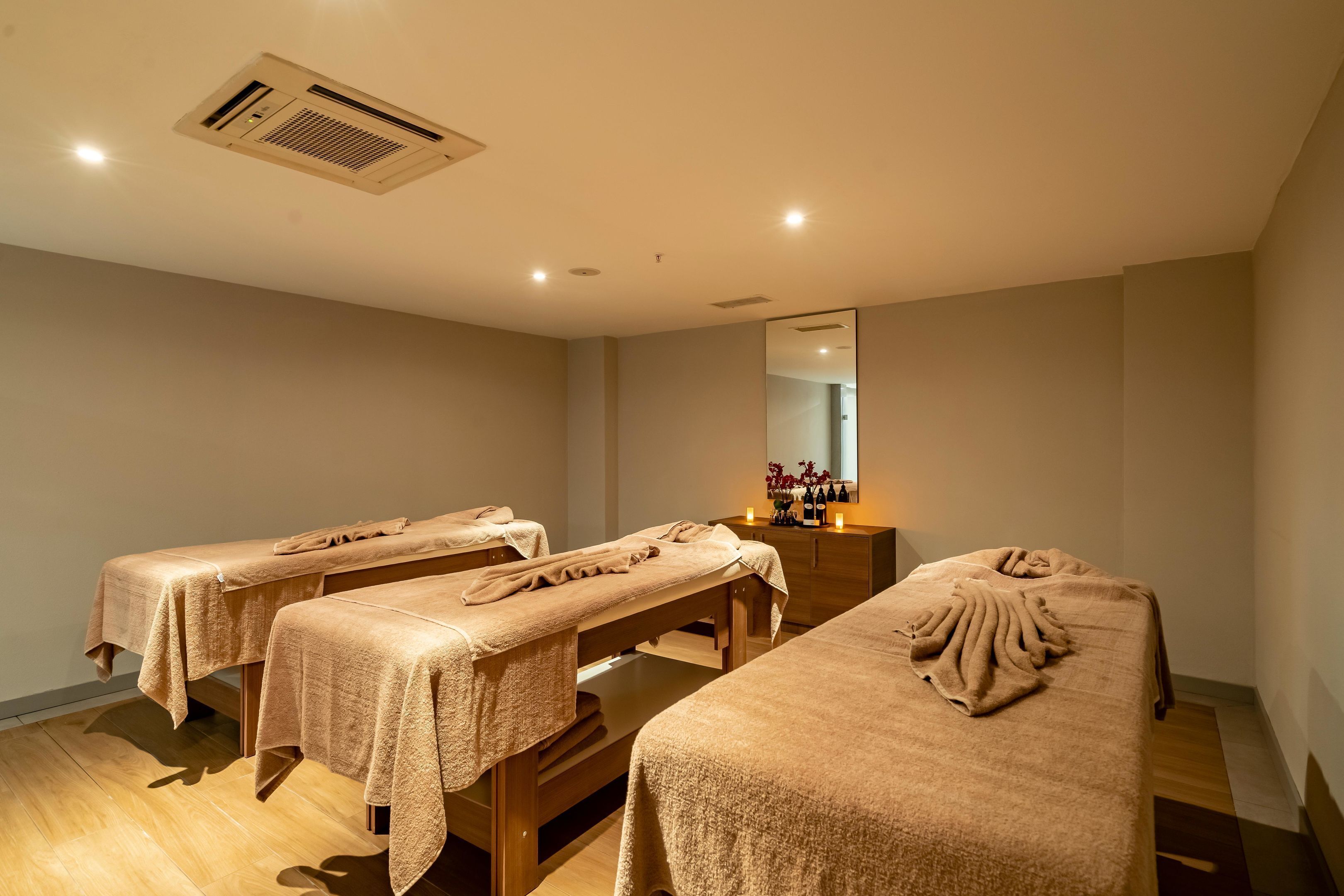 Ein Raum mit drei Massagetischen, Holzboden, einem Deckenlüftungsgitter und einem Spiegel an der Wand.