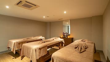 Ein Raum mit drei Massagetischen, Holzboden, einem Deckenlüftungsgitter und einem Spiegel an der Wand.