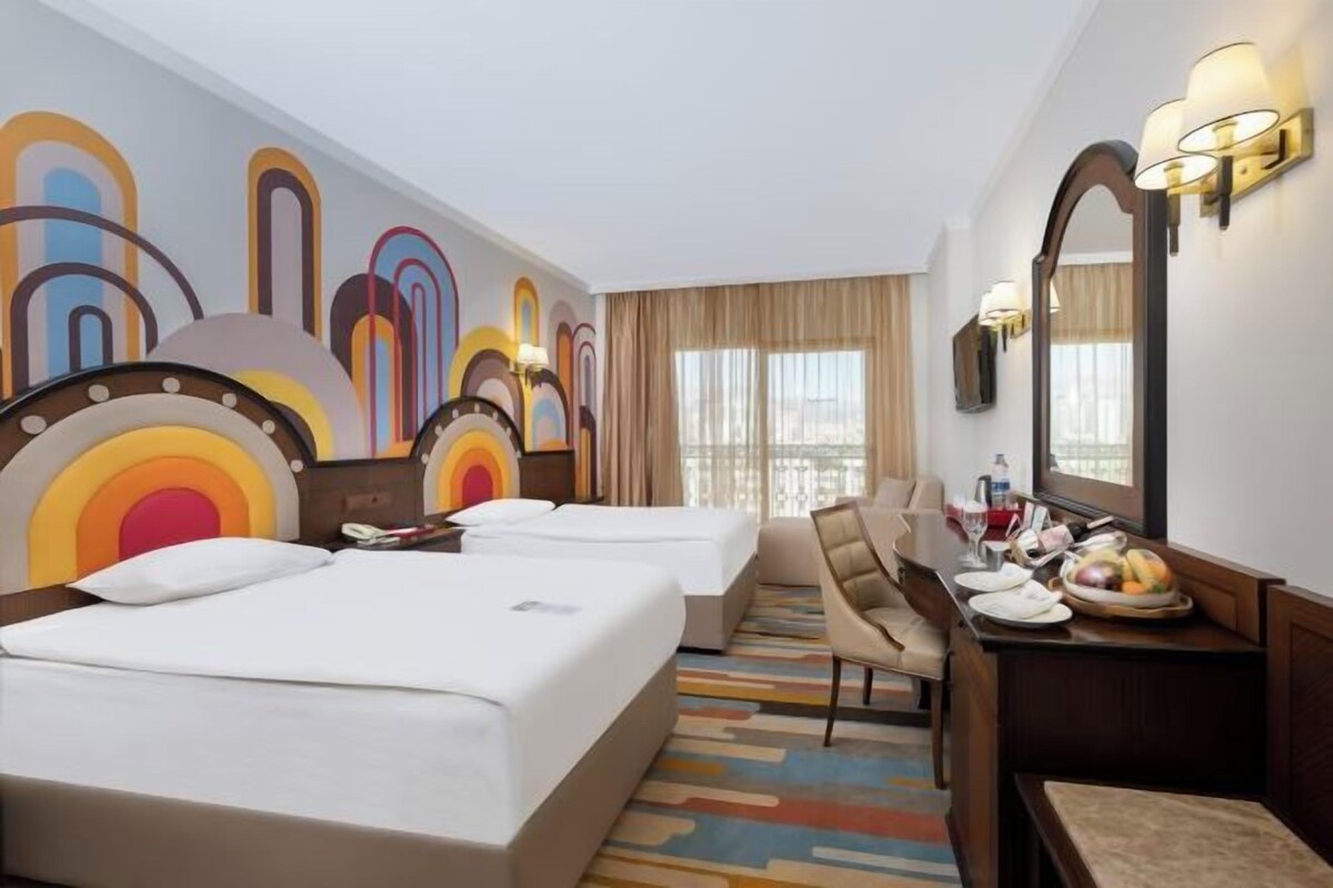 A hotel room with two beds, a desk with a chair, a dining table with a fruit bowl, and a large window with curtains.