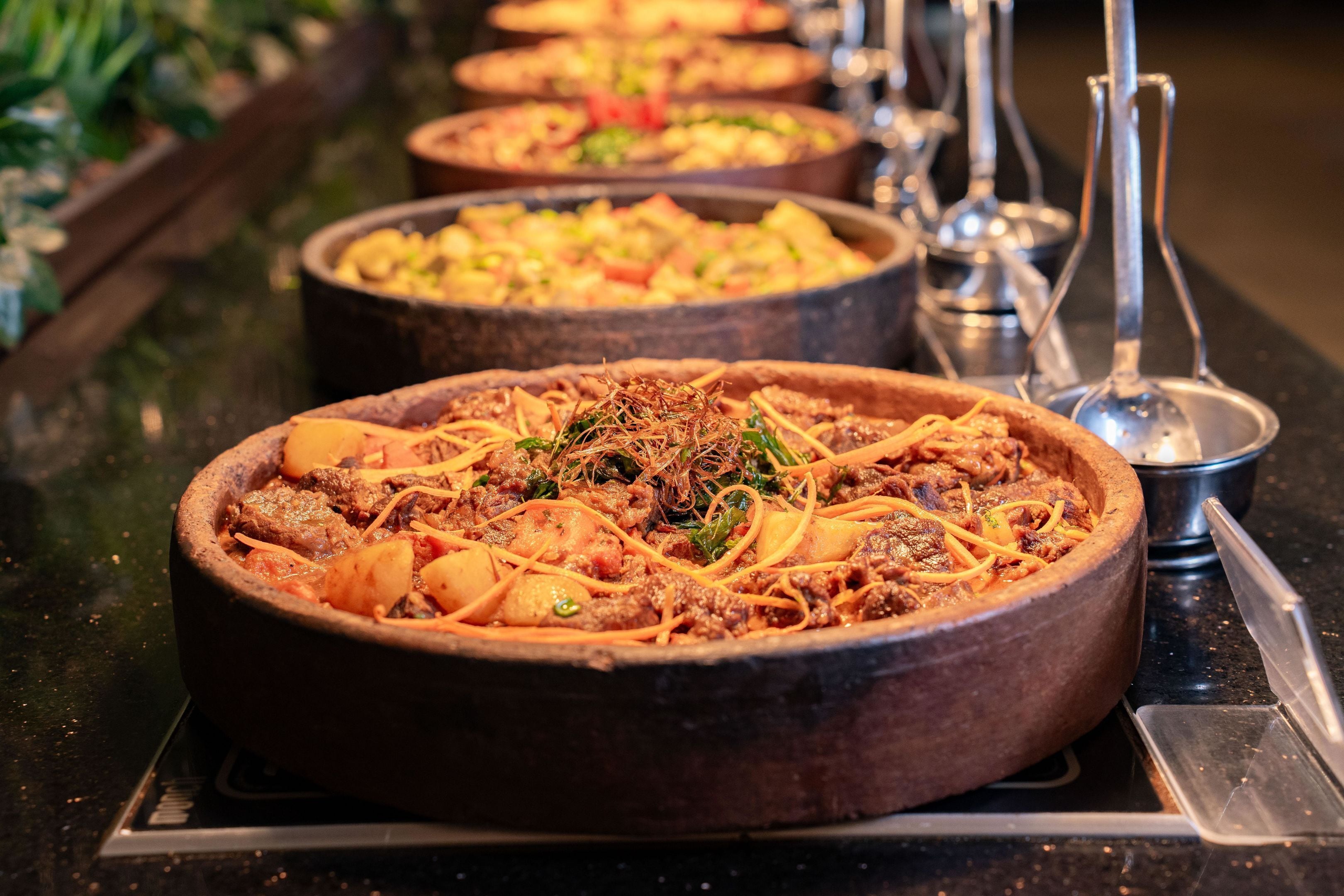 Un bufé con varios platos en cuencos de madera, incluyendo un plato de carne con verduras y guarniciones.