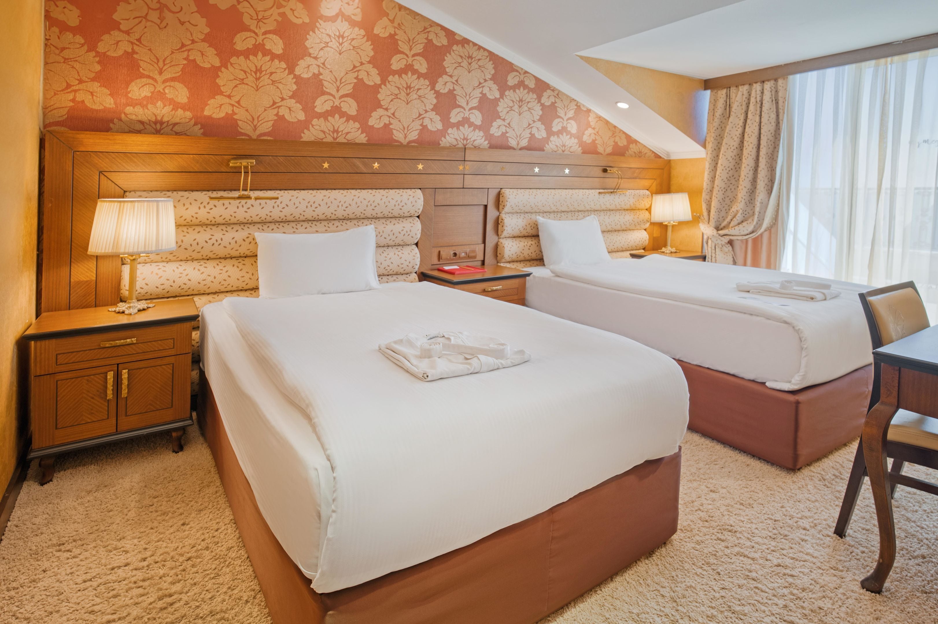 A hotel room with two beds, a wooden headboard, a nightstand with a lamp, a wooden dresser, and a window with curtains.