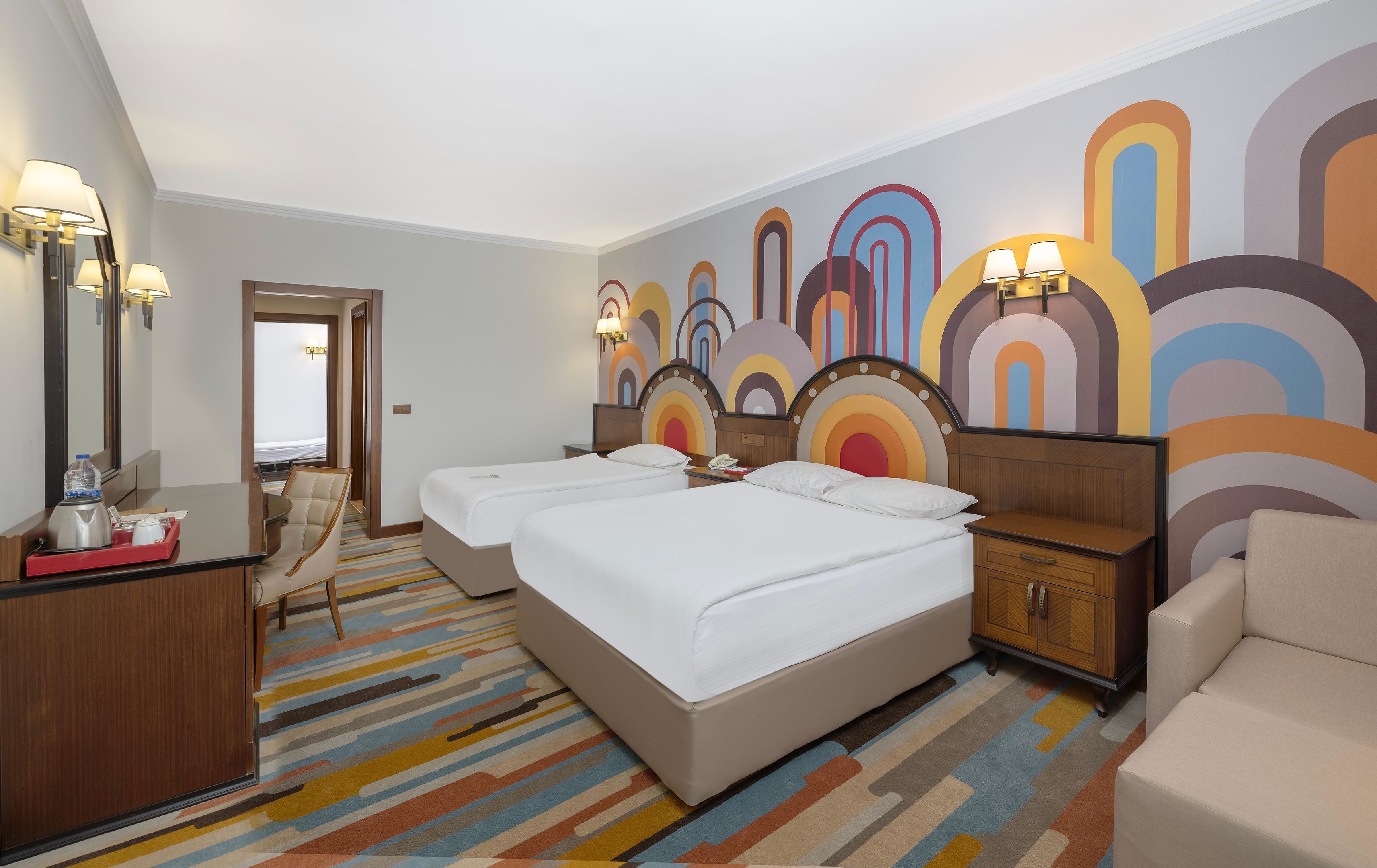 A hotel room with two beds, a desk with a mirror, a chair, a nightstand, and a colorful abstract wall mural.