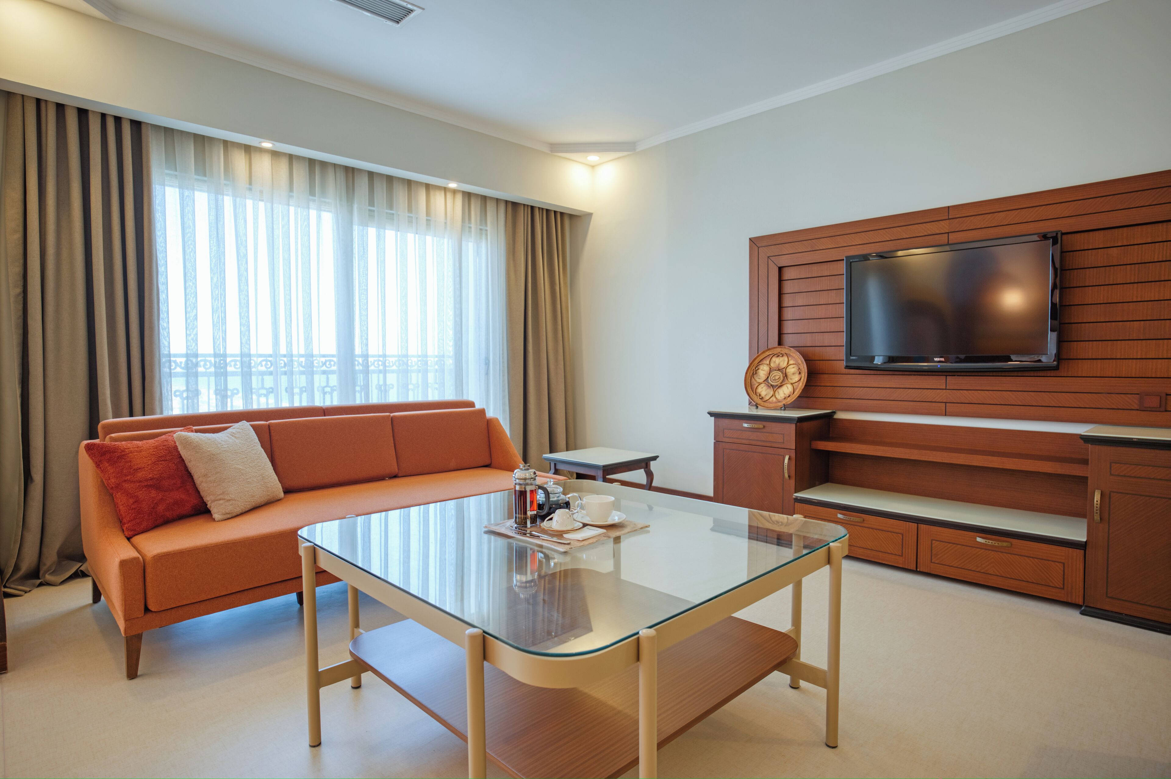 A modern hotel room with a glass-top table, orange sofa, wooden TV stand, and a large window with curtains.