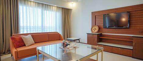 A modern hotel room with a glass-top table, orange sofa, wooden TV stand, and a large window with curtains.