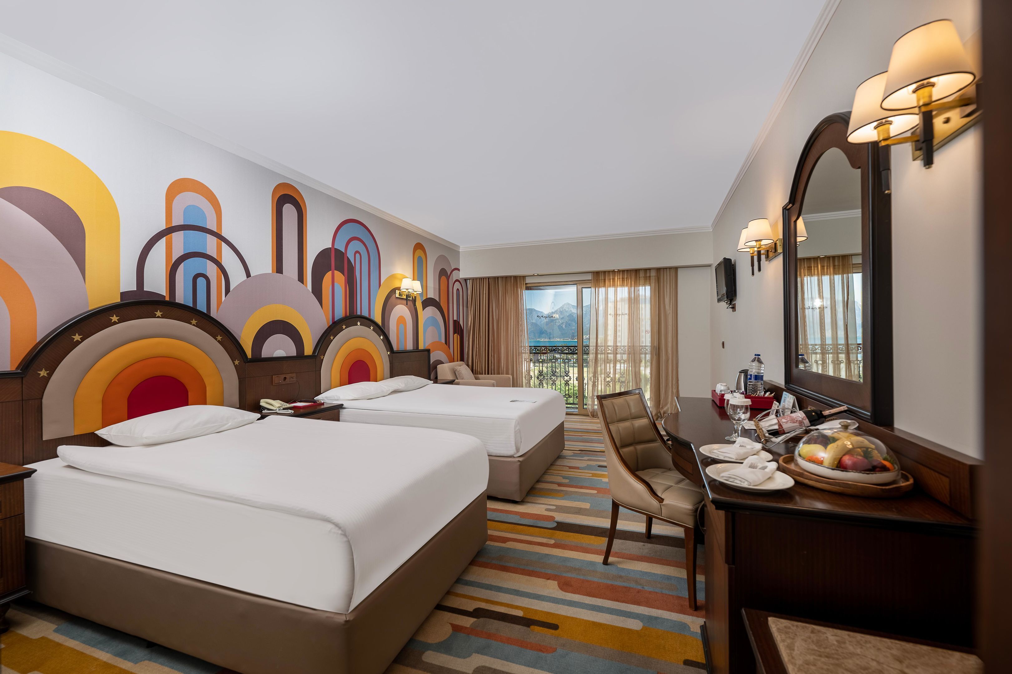 A hotel room with two beds, a dining table with a fruit bowl, a mirror, and a view of mountains through the window.