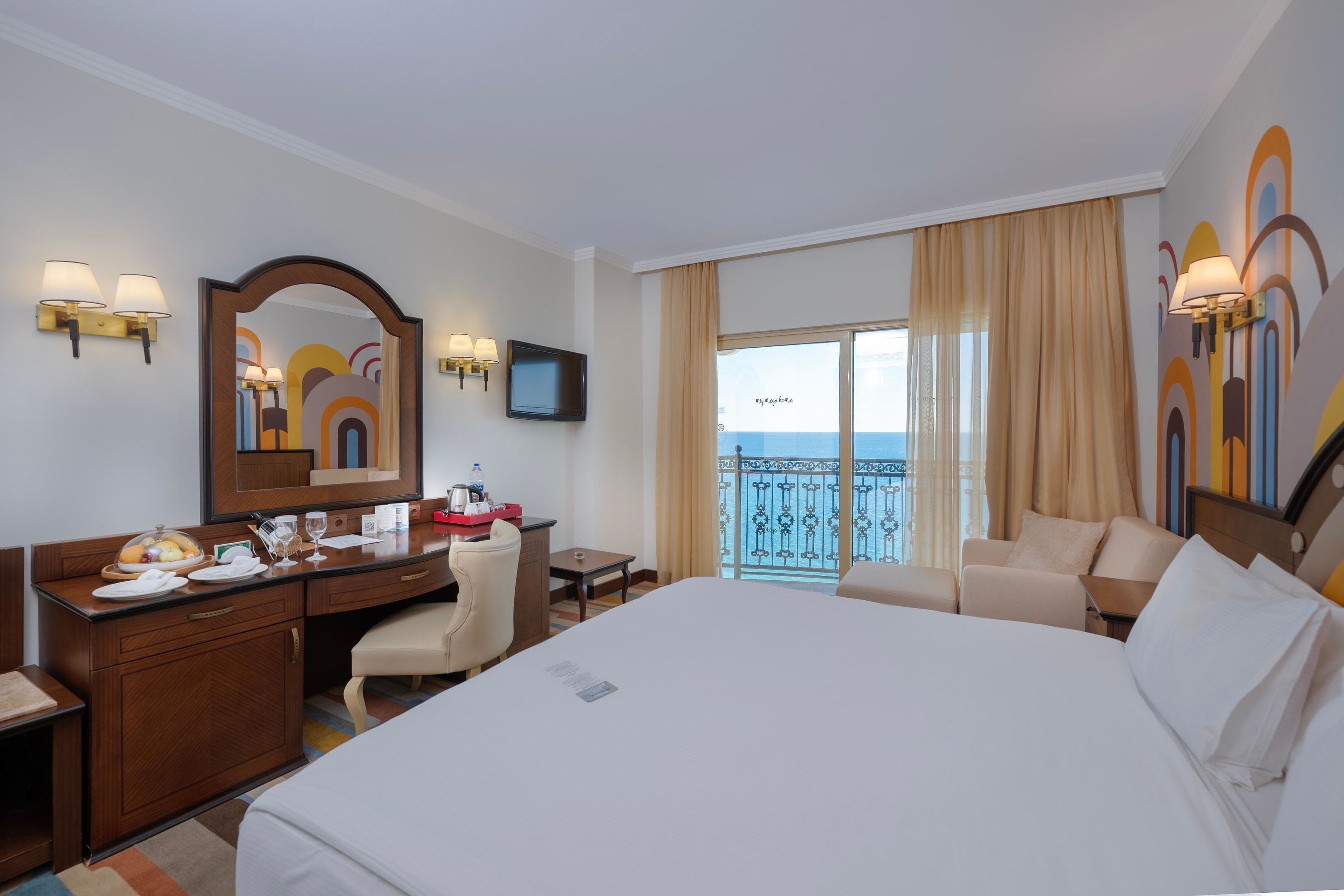 Ein Hotelzimmer mit einem großen Bett, einem Schreibtisch mit Stuhl, einem kleinen Tisch, einem Spiegel und einem Fernseher. Es gibt einen Balkon mit Meerblick.