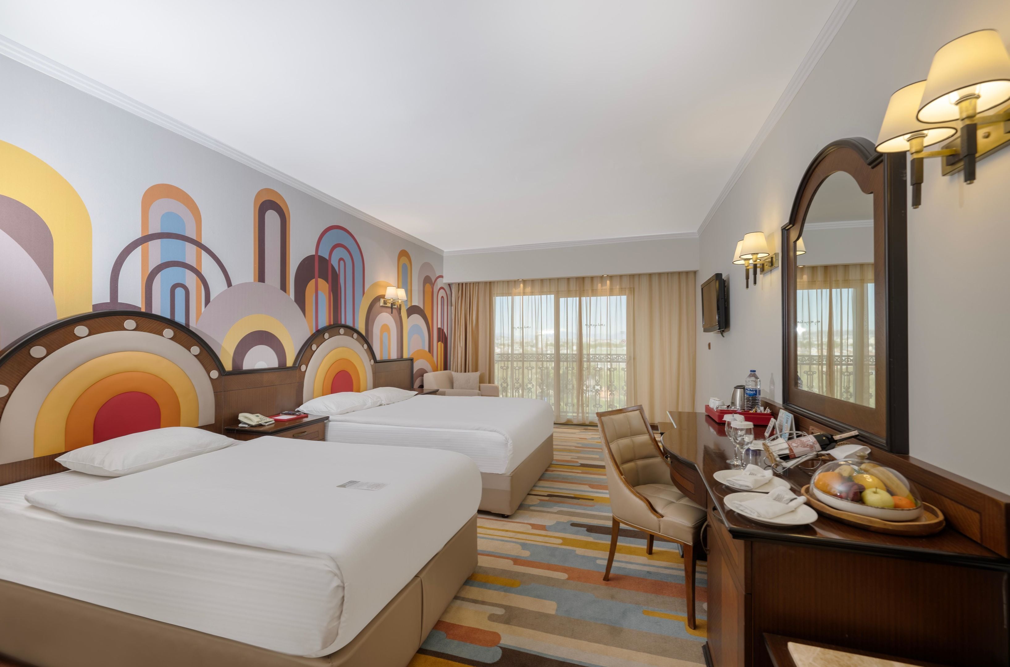 A hotel room with three beds, a desk with a chair, a dining table with a fruit bowl, and a large mirror.