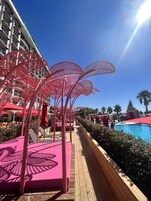 A resort pool area with red palm-like structures, lounge chairs, and a clear blue sky.