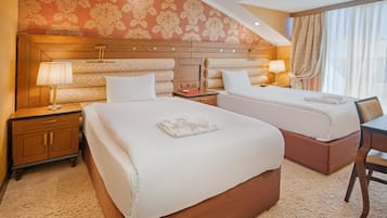 Habitación de hotel con dos camas, cabecero de madera, mesita de noche con lámpara, cómoda de madera y ventana con cortinas.