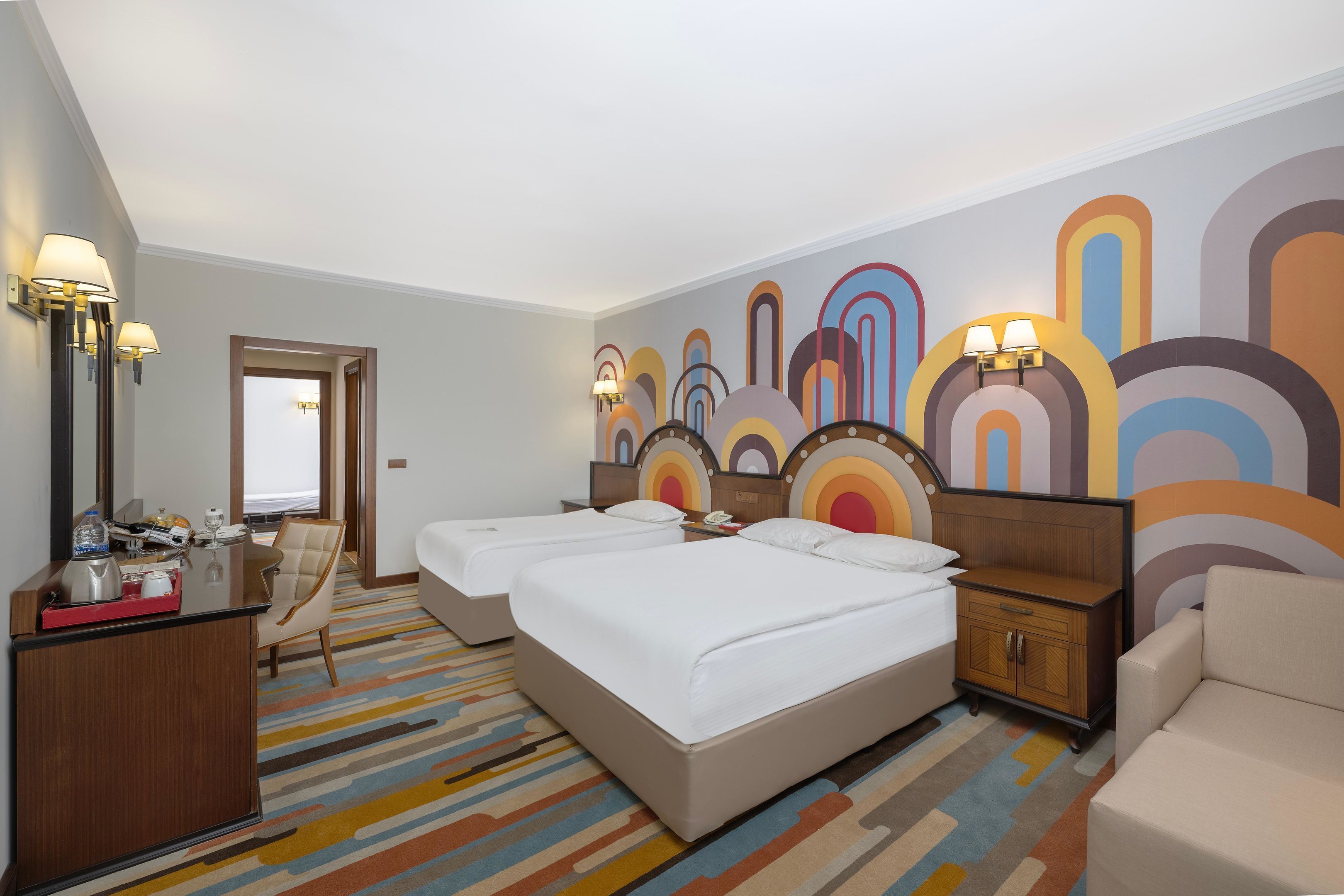 Une chambre d’hôtel avec deux lits, un bureau avec une chaise, une commode et une fresque murale abstraite aux couleurs vives.