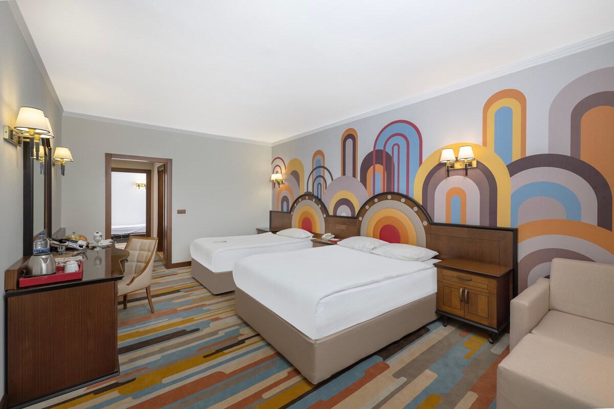 A hotel room with two beds, a desk with a chair, a dresser, and a colorful abstract wall mural.